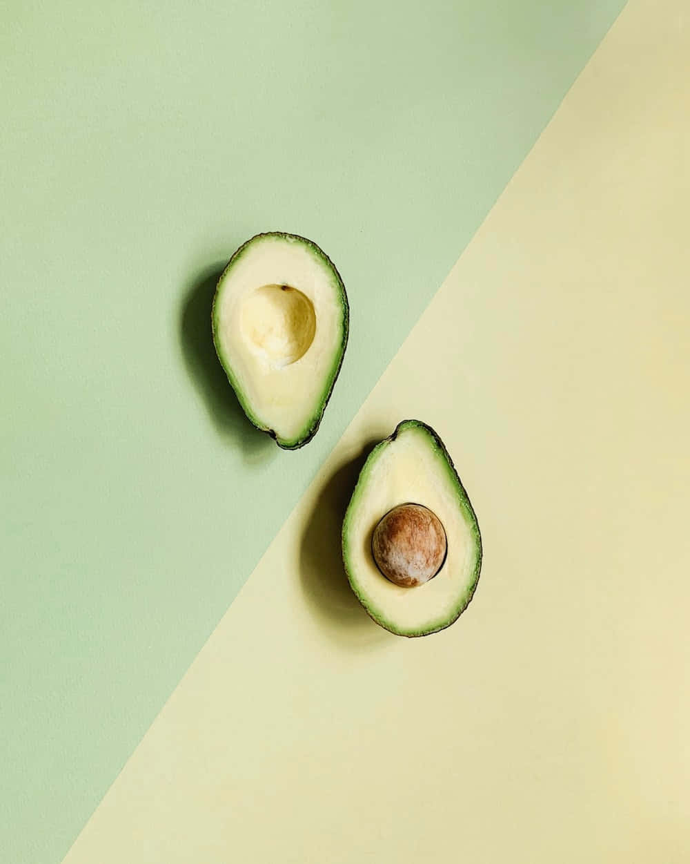 Avocado 桌布 壁紙 — 兩個牛油果半邊放在一個分為綠色和米色的背景上，一個有棕色果核，另一個空的。