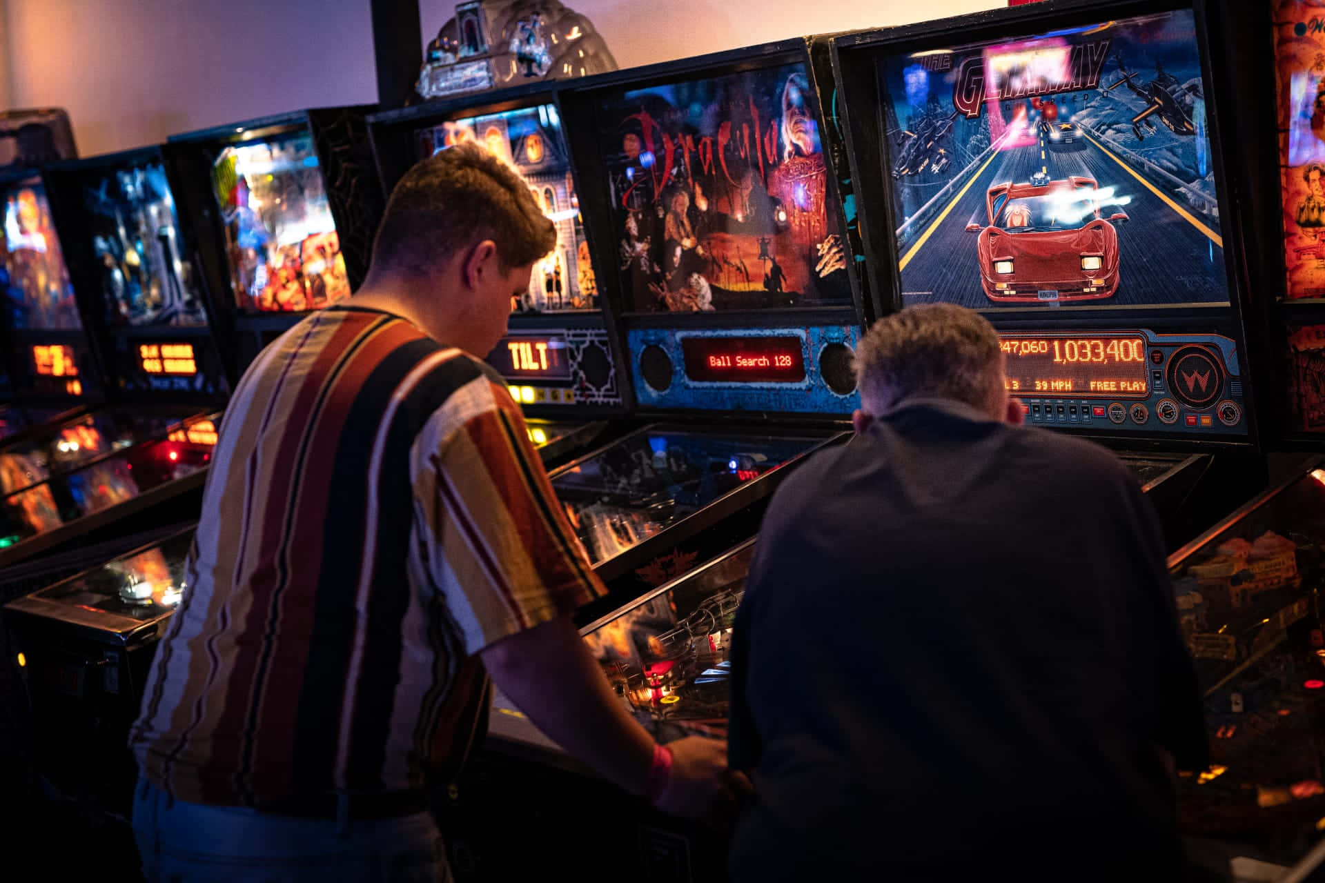 Arcade Games 桌布 壁紙 — 兩個男人在昏暗的遊戲室中，在閃爍的街機燈光下激烈地玩著復古彈珠機。