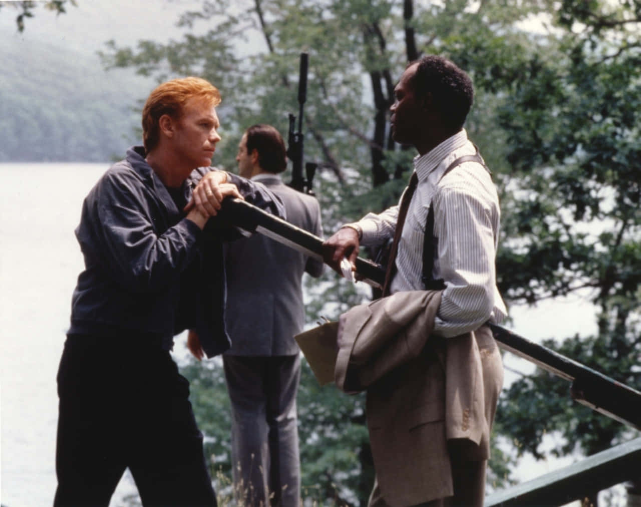 David Caruso 桌布 壁紙 — 兩個男人在湖邊碼頭上為一支步槍對峙，身後有樹木和水面。