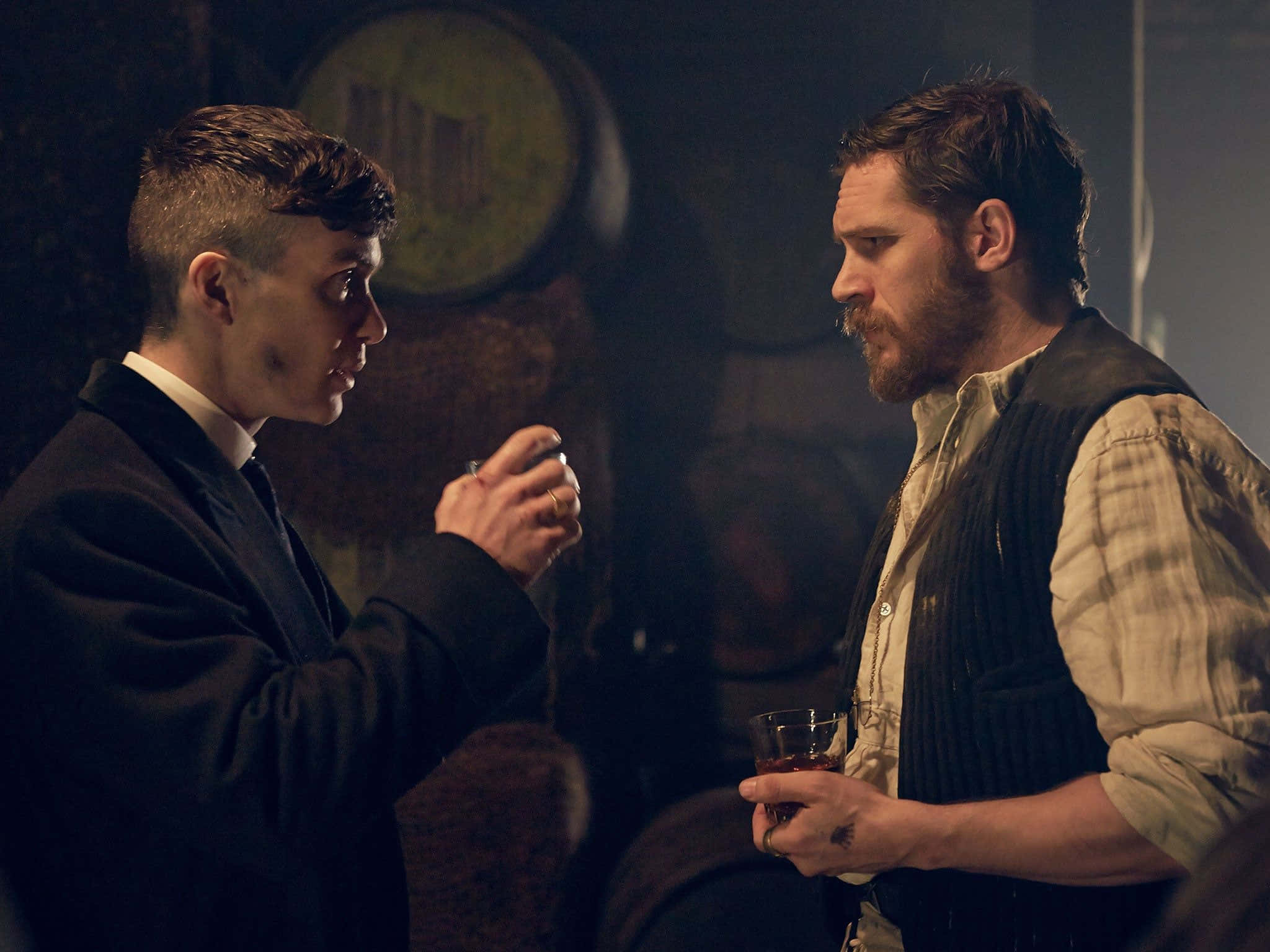 Peaky Blinders 桌布 壁紙 — 兩個男人在燈光昏暗的酒吧裡對峙，表情緊張，一個拿著酒杯，另一個用手指上的戒指做手勢。