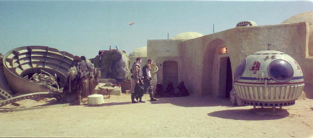 Mos Eisley 桌布 壁紙 — 兩個男人站在沙漠住所前，附近有一艘失事的太空船和一個球形車輛，天空清澈湛藍。
