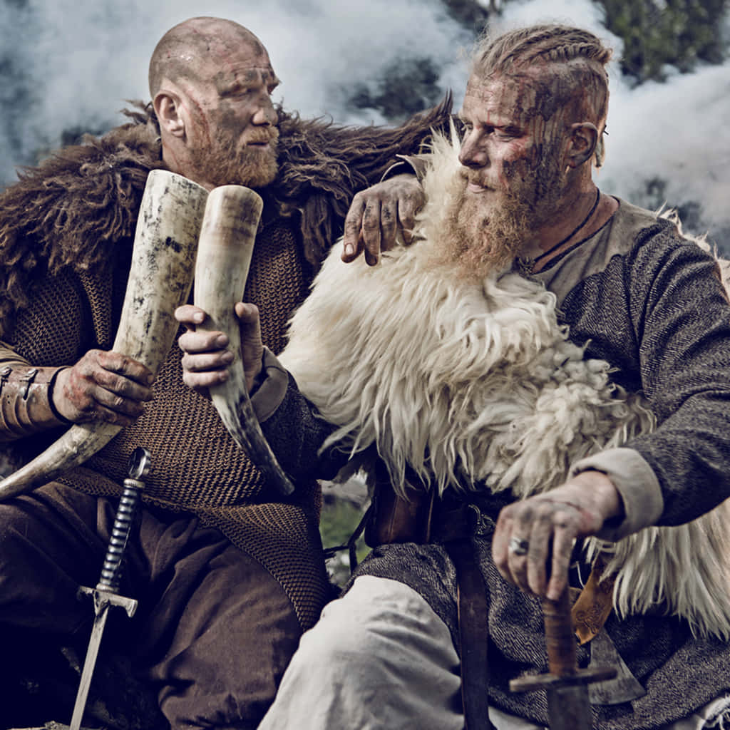 Vikings 桌布 壁紙 — 兩個留鬍子的維京人身穿毛皮襯裏盔甲並排而坐，一個手持角，另一個緊握白色毛皮，身邊放著劍。