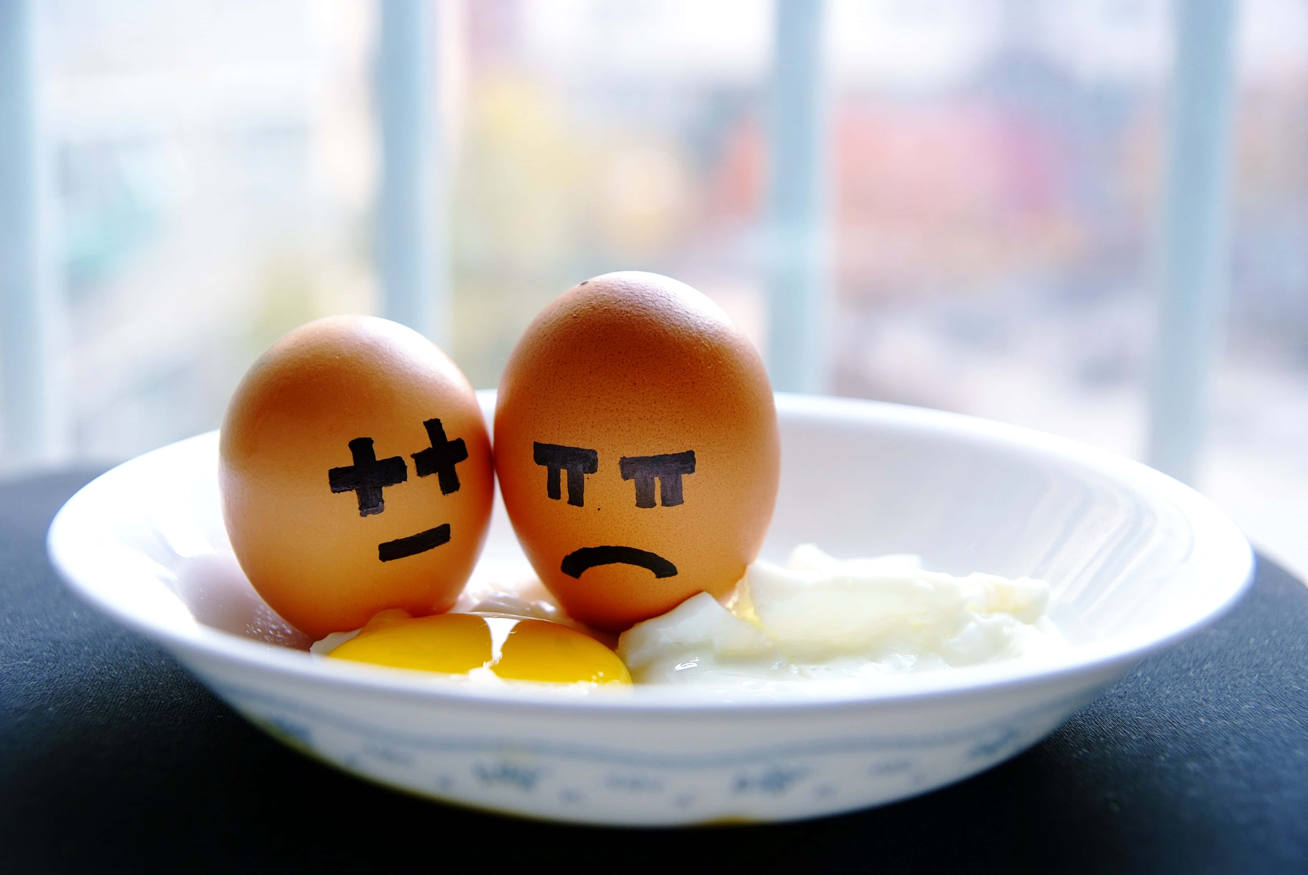 Brown Eggs 桌布 壁紙 — 兩個畫上憤怒表情的棕色雞蛋放在白色碗中，旁邊是破裂的蛋黃和蛋白，位於窗邊的深色桌子上。