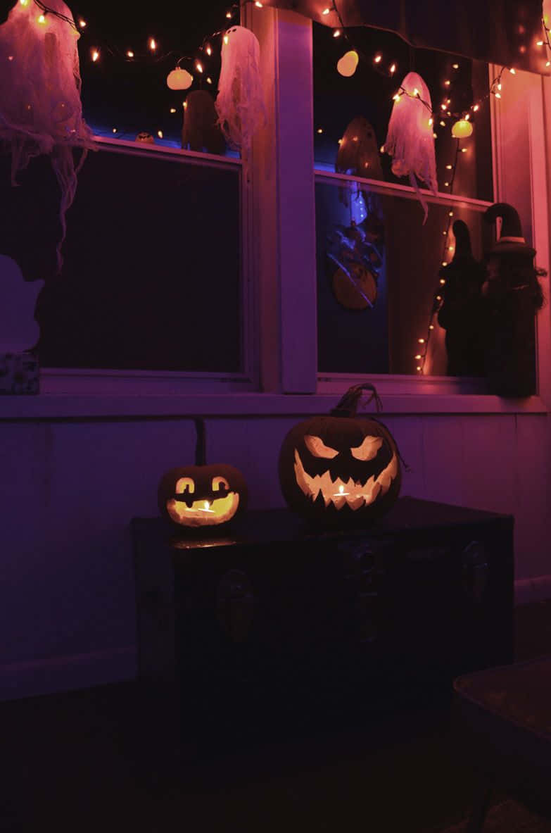 Halloweentown 桌布 壁紙 — 兩個發光的南瓜燈放在一個行李箱上，下方有紫色串燈，窗邊掛著鬼魂裝飾，夜晚時分。