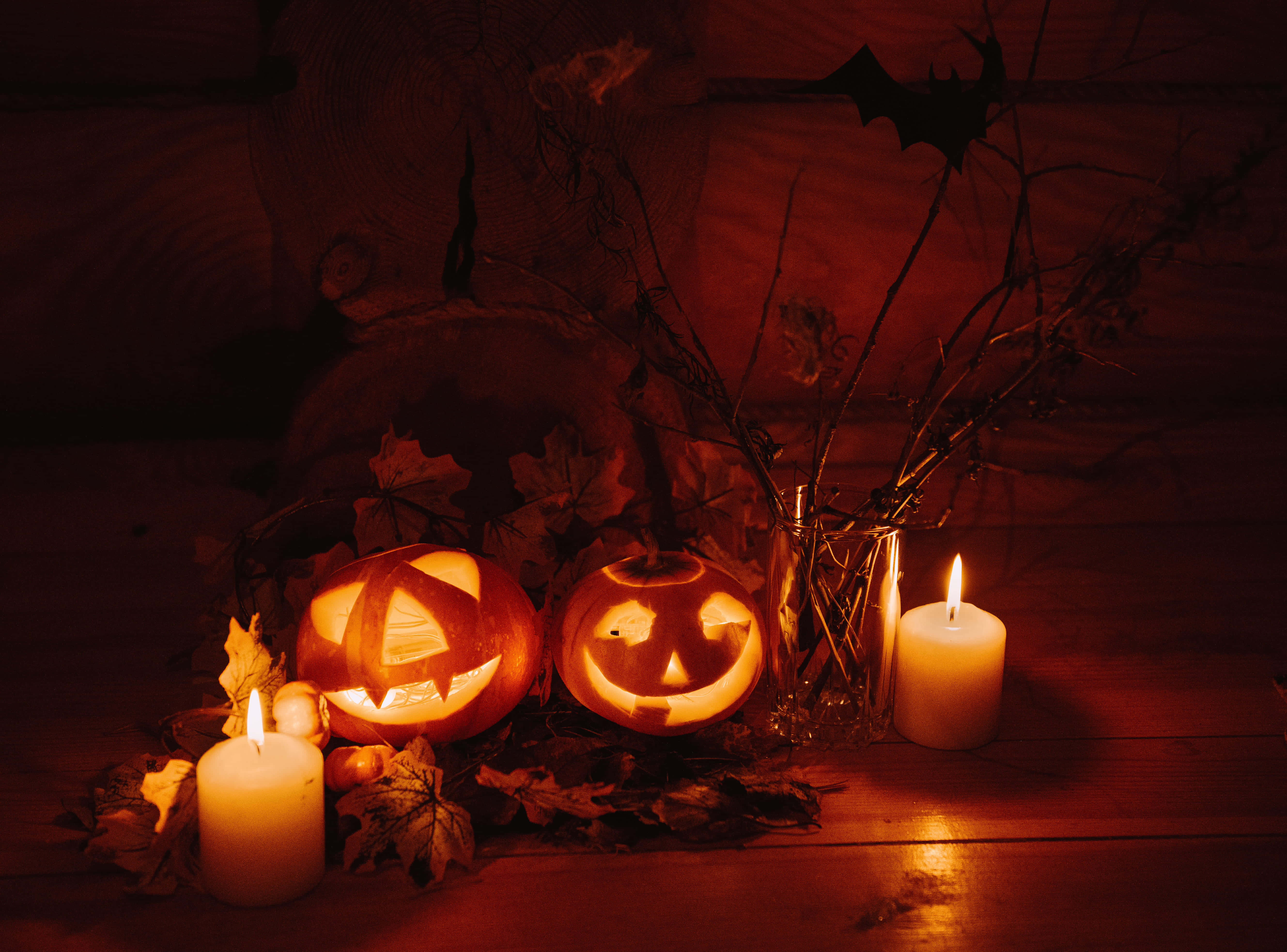 Halloween Candles 桌布 壁紙 — 兩個發光的南瓜燈籠，蠟燭在木質背景上為秋葉和蝙蝠剪影投射溫暖的光芒。