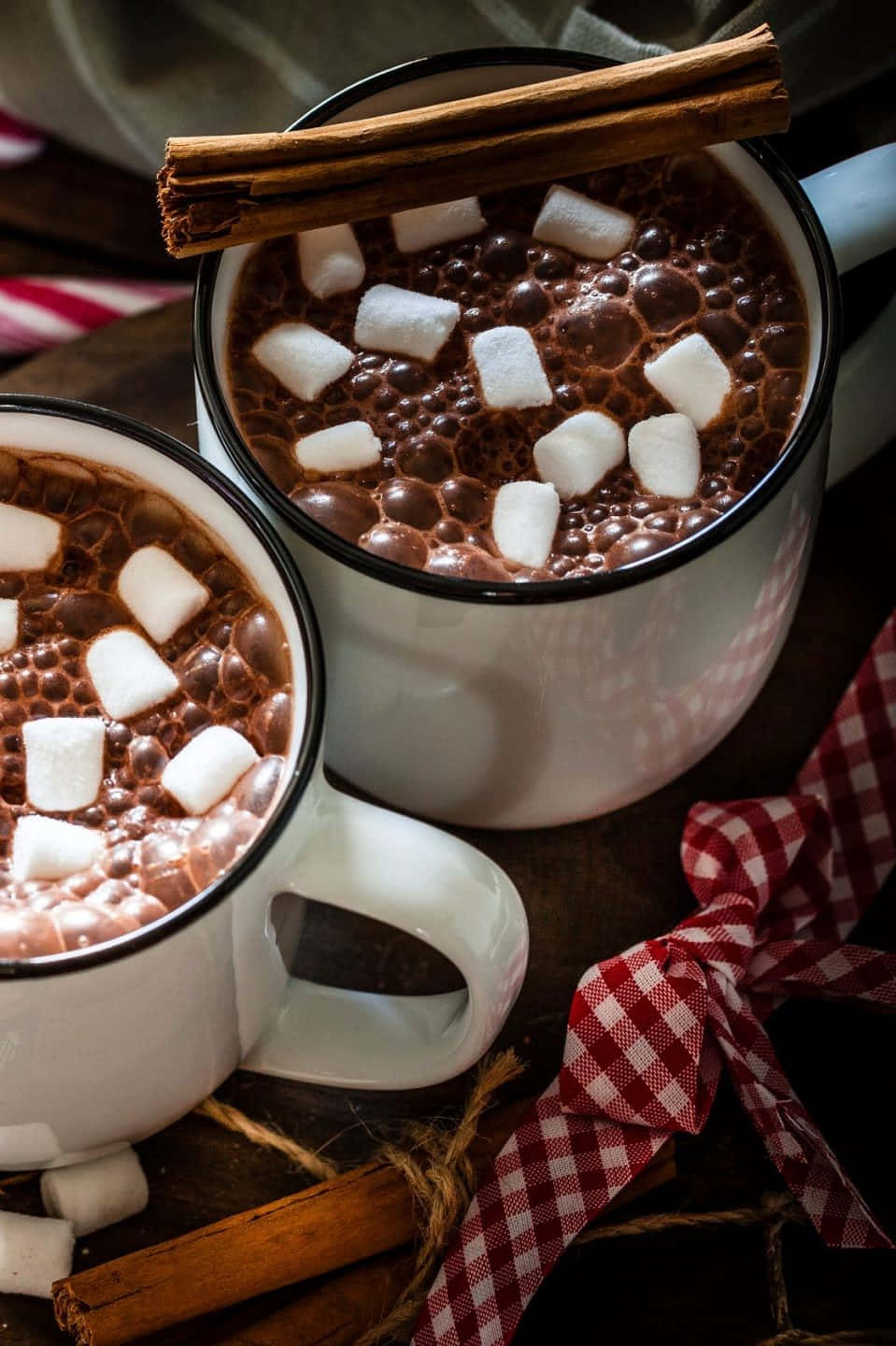 Hot Chocolate 桌布 壁紙 — 兩個白色琺瑯杯，裝滿冒著熱氣的熱朱古力，上面放有棉花糖和肉桂棒，置於鄉村風木桌上。
