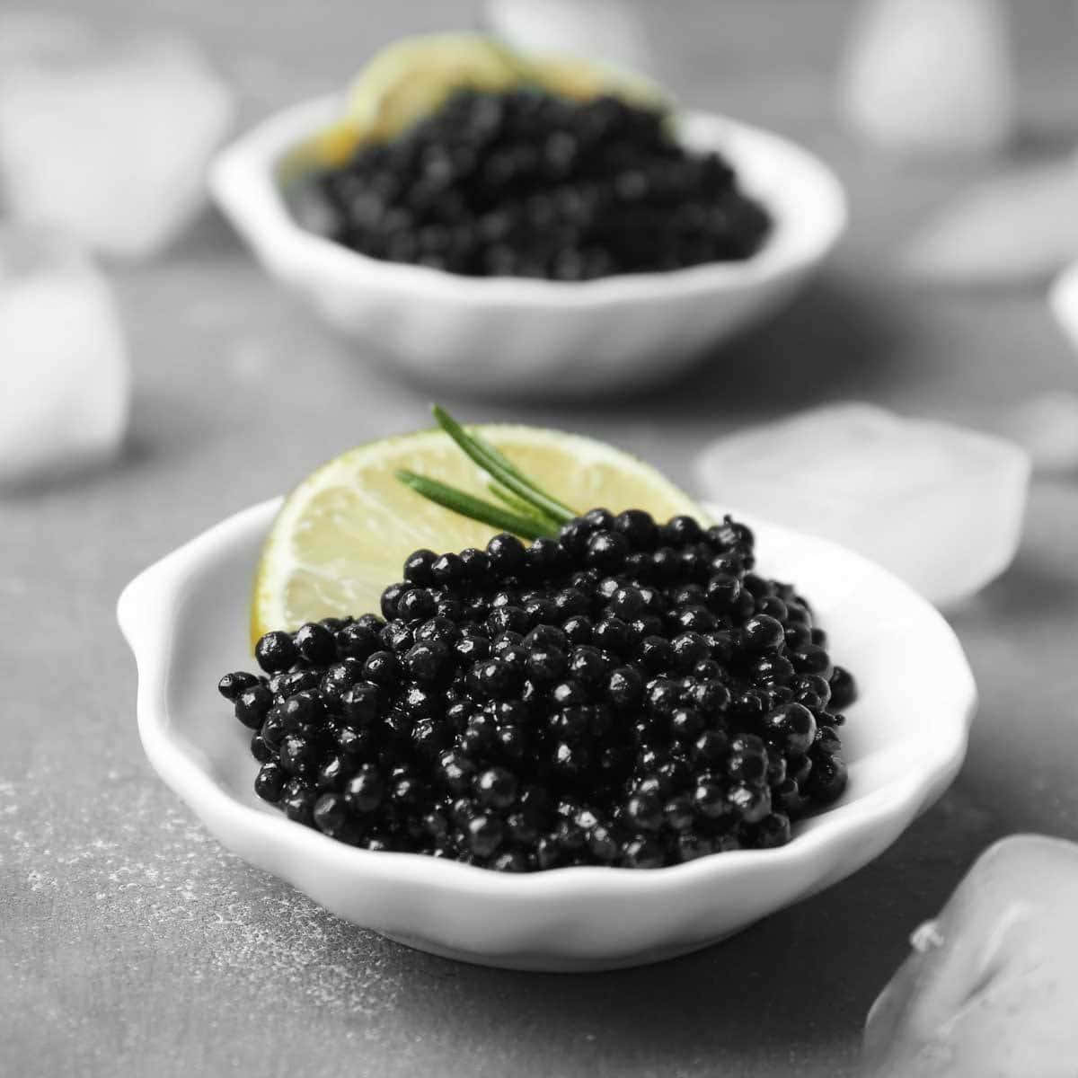 Black Caviar 桌布 壁紙 — 兩個白色碗裝滿黑魚子醬，配以檸檬和迷迭香裝飾，放在被冰塊包圍的灰色表面上。