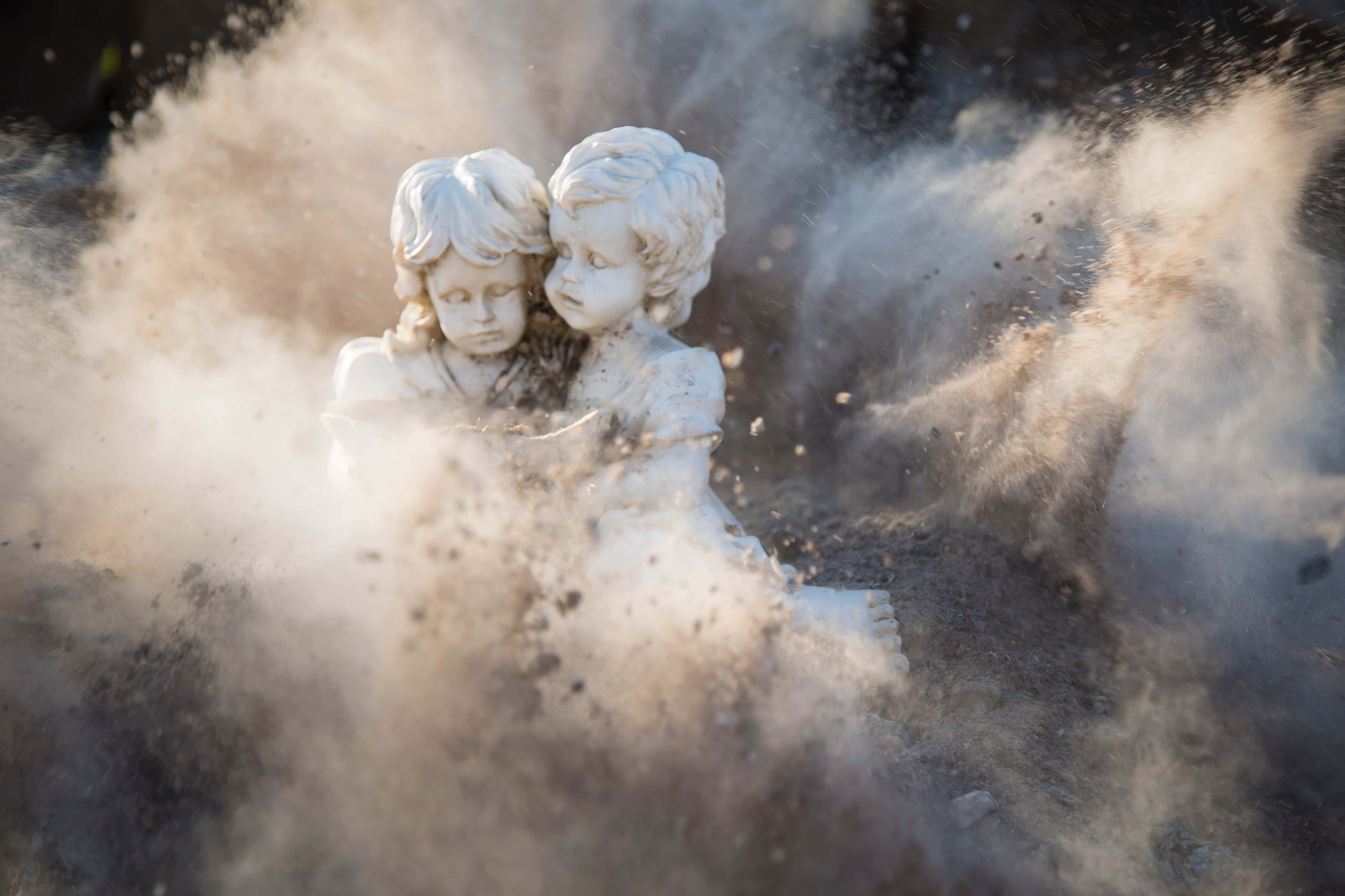 Dust 桌布 壁紙 — 兩個石製小天使雕像擁抱，灰塵與碎片在它們周圍以戲劇性的慢動作爆炸。