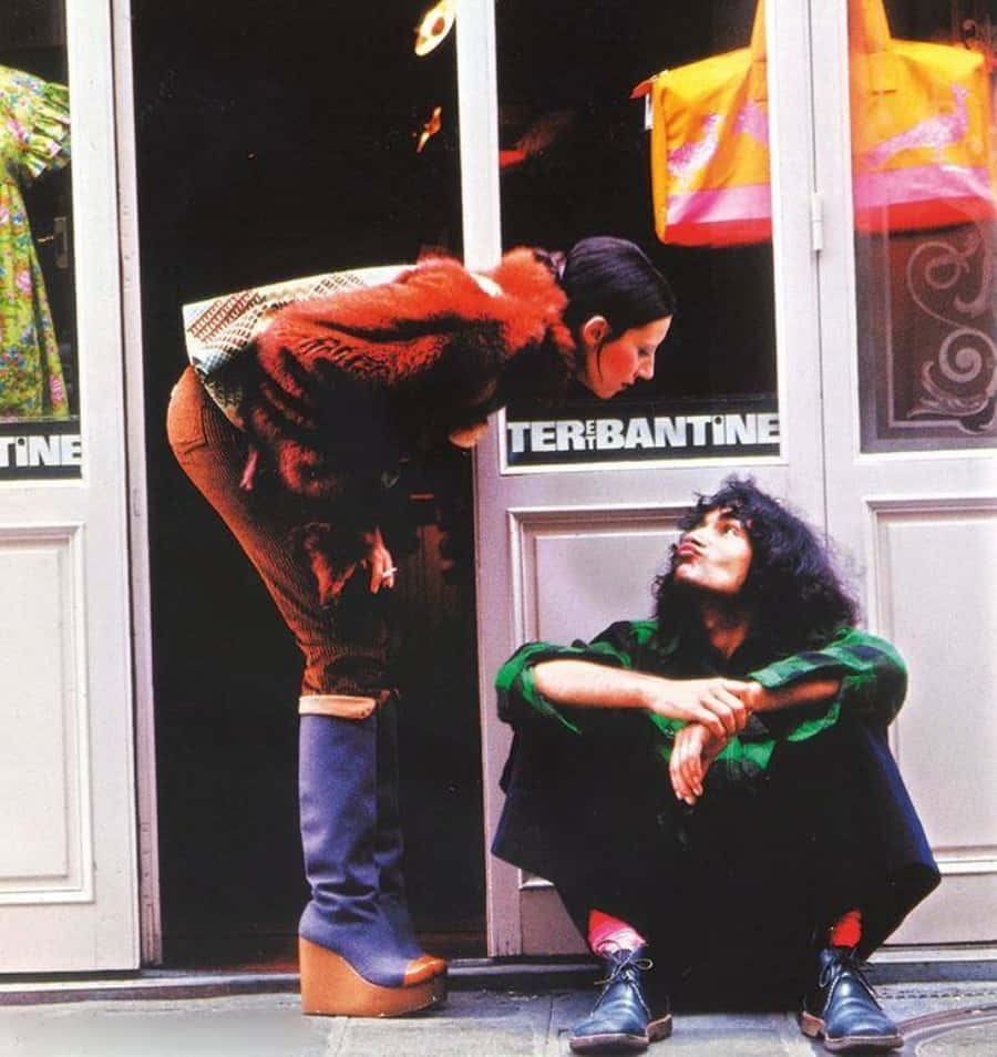 70年代gif 桌布 壁紙 — 兩個穿著1970年代時尚的人在商店門附近互動，一人蹲下，另一人彎腰穿著厚底靴和毛領。