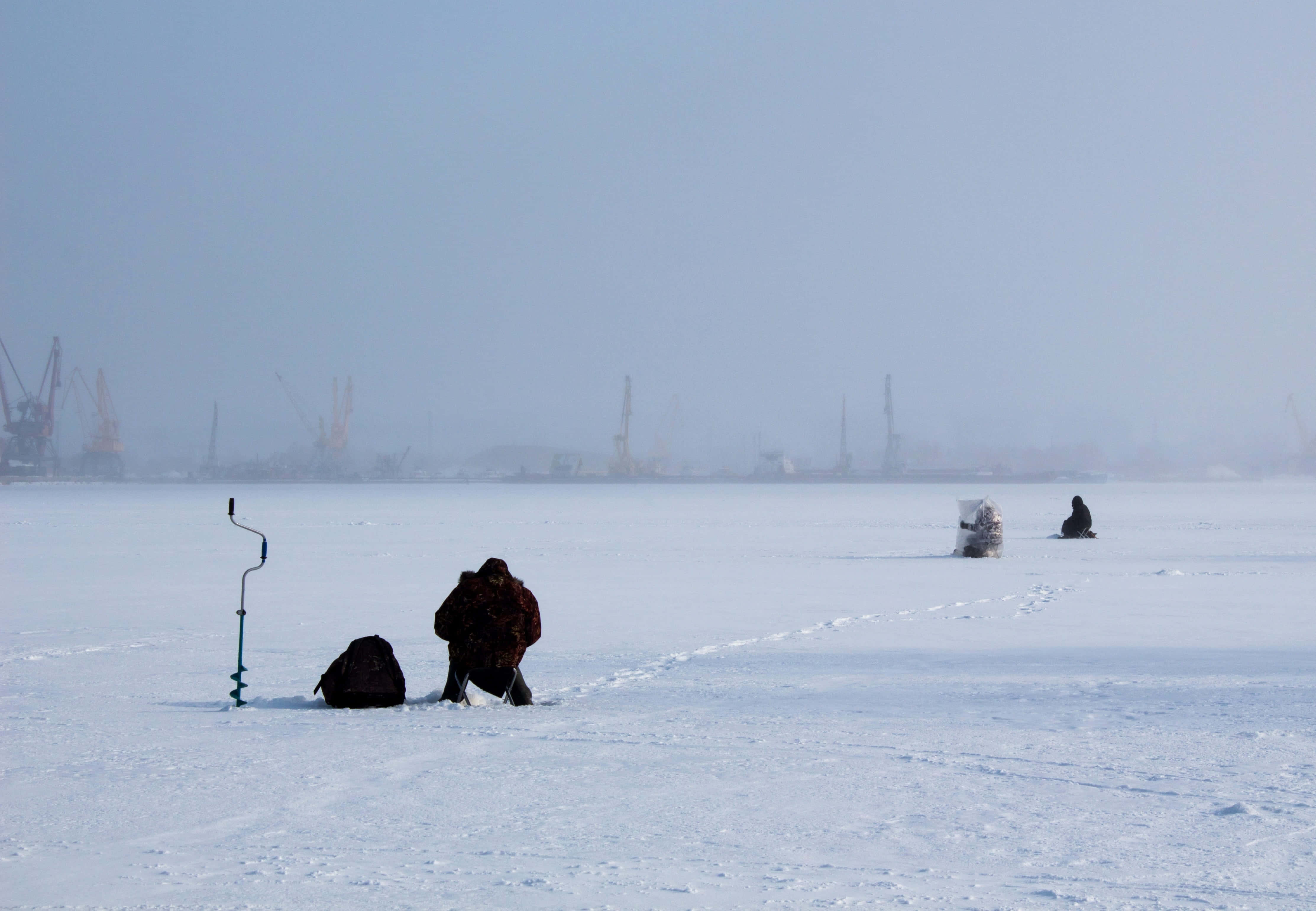 Ice Fishing 桌布 壁紙 — 兩個穿著厚重冬季裝備的人物在廣闊的冰凍湖面上鑽孔，遠處隱約可見工業起重機。