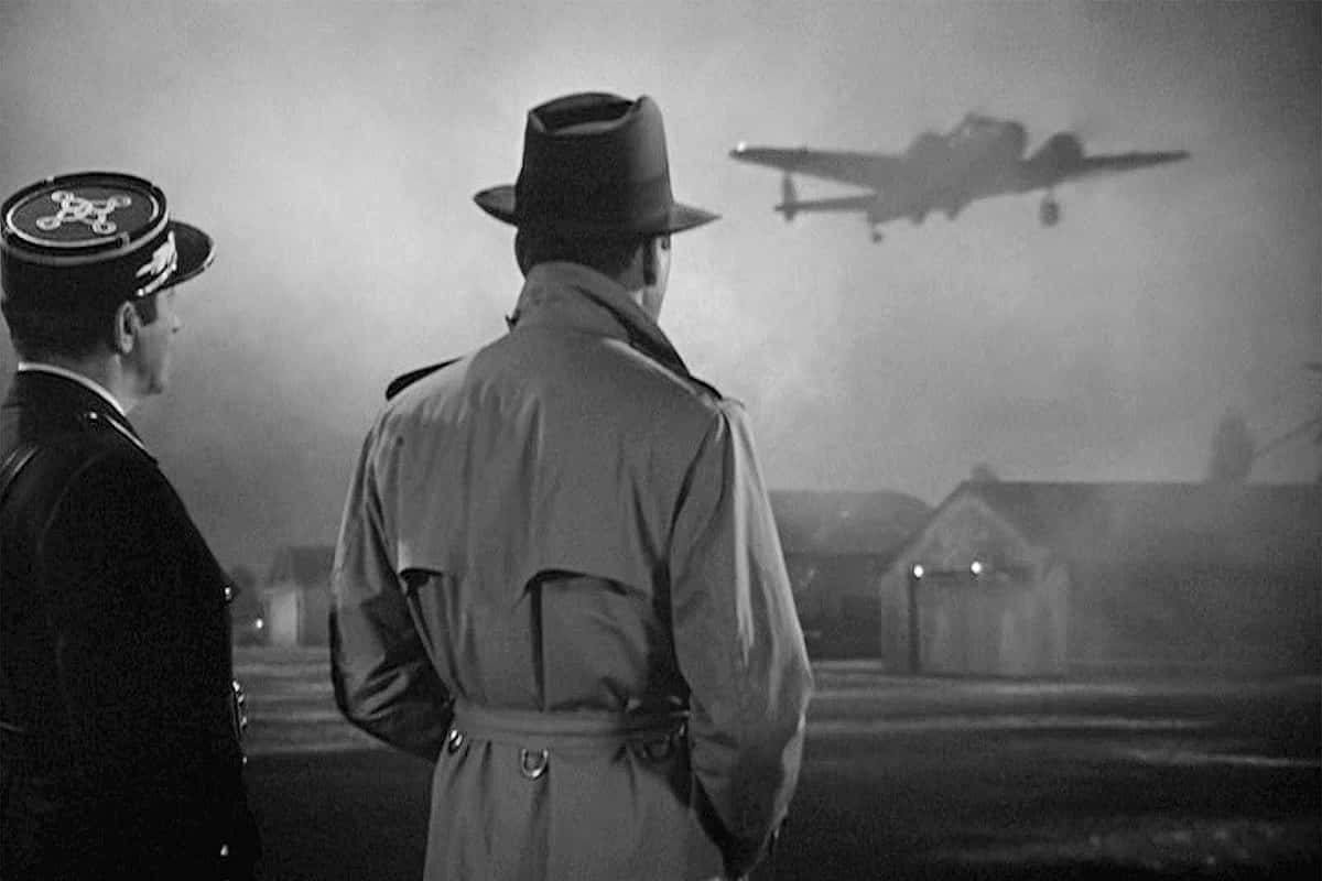 Film Noir 桌布 壁紙 — 兩個穿著大衣和戴帽子的男人看著一架復古飛機在黃昏時低飛過霧氣瀰漫的鄉村景觀。