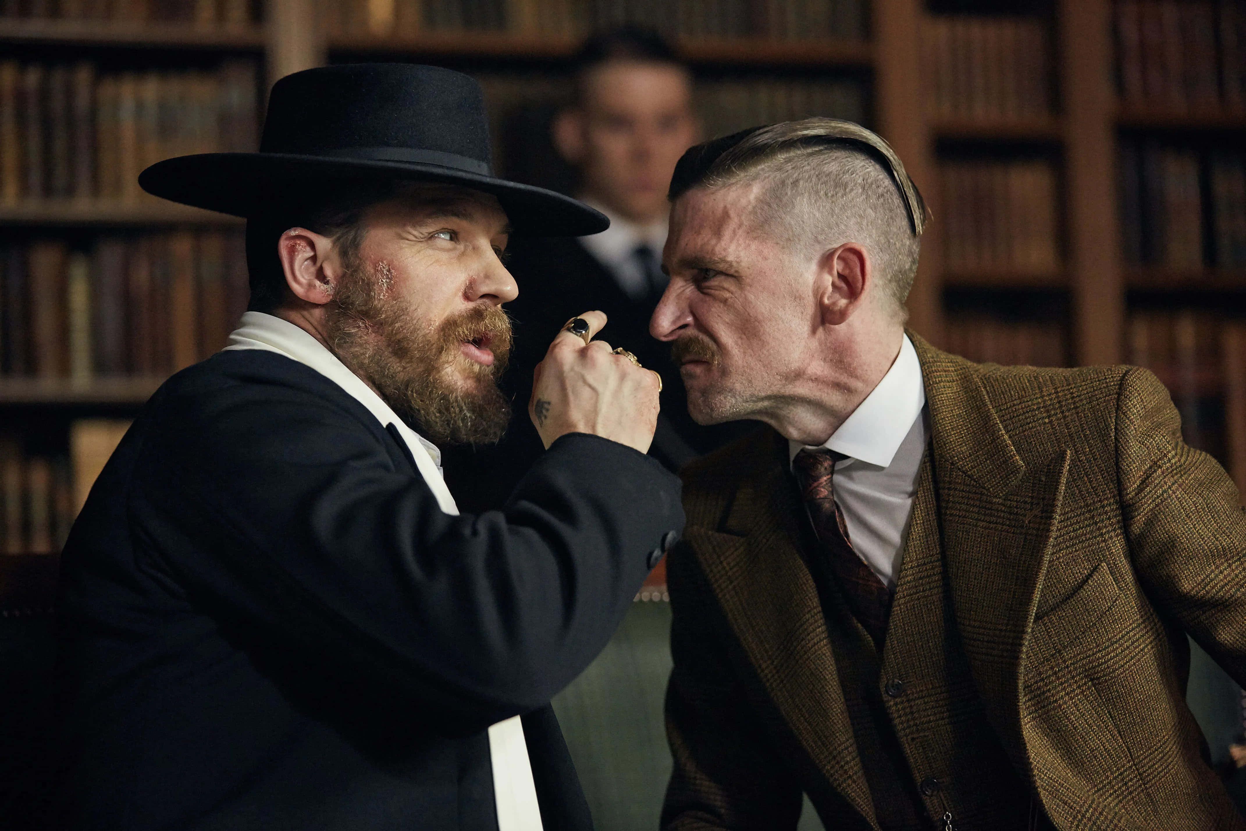 Peaky Blinders 桌布 壁紙 — 兩個穿著復古西裝的男人在一個光線昏暗、充滿書架的圖書館中激烈對峙。