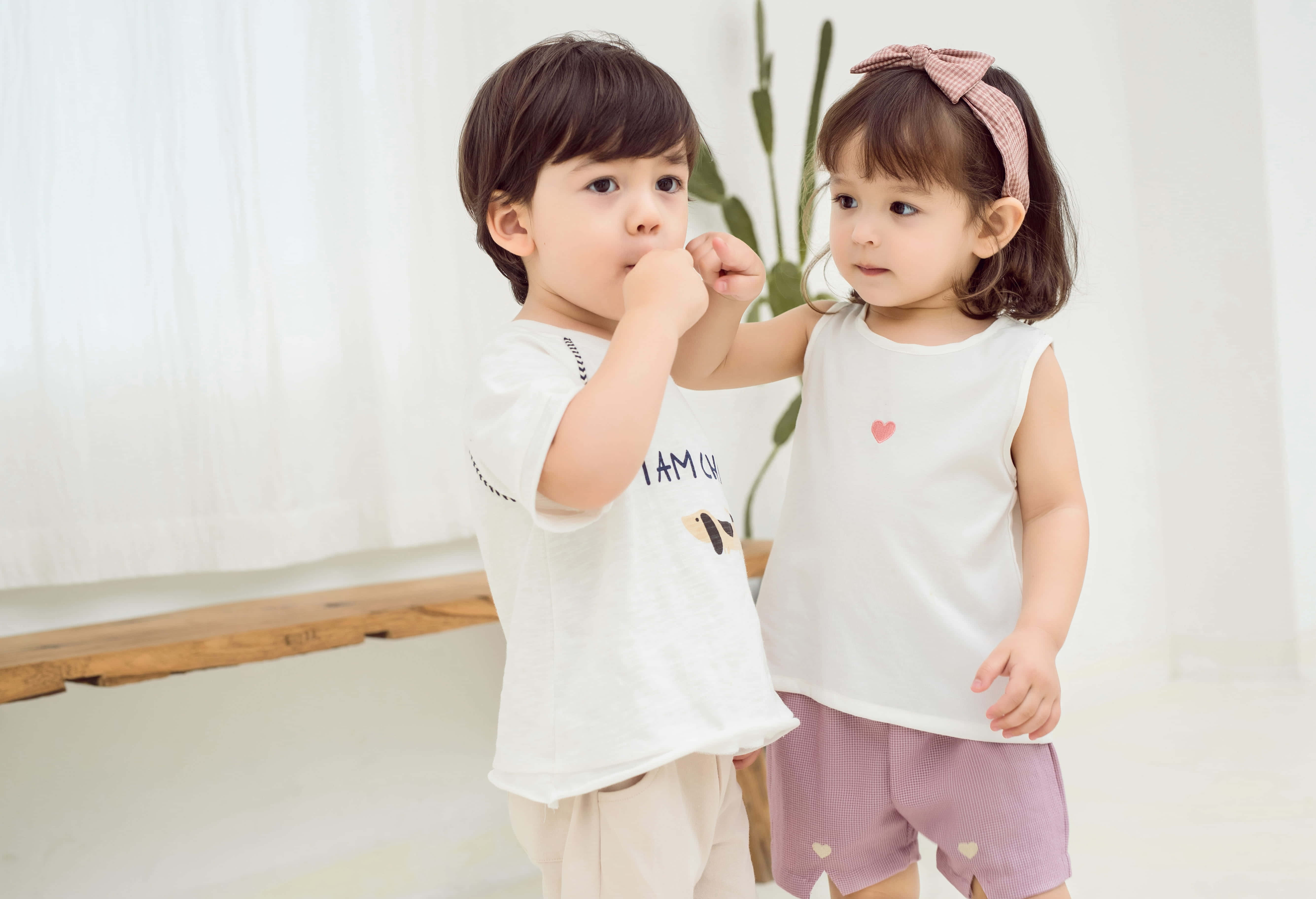 Cute Kids 桌布 壁紙 — 兩個穿著白色衣服的幼兒在室內靠近站立，一個咬手指，另一個戴著粉紅色蝴蝶結頭帶看著。