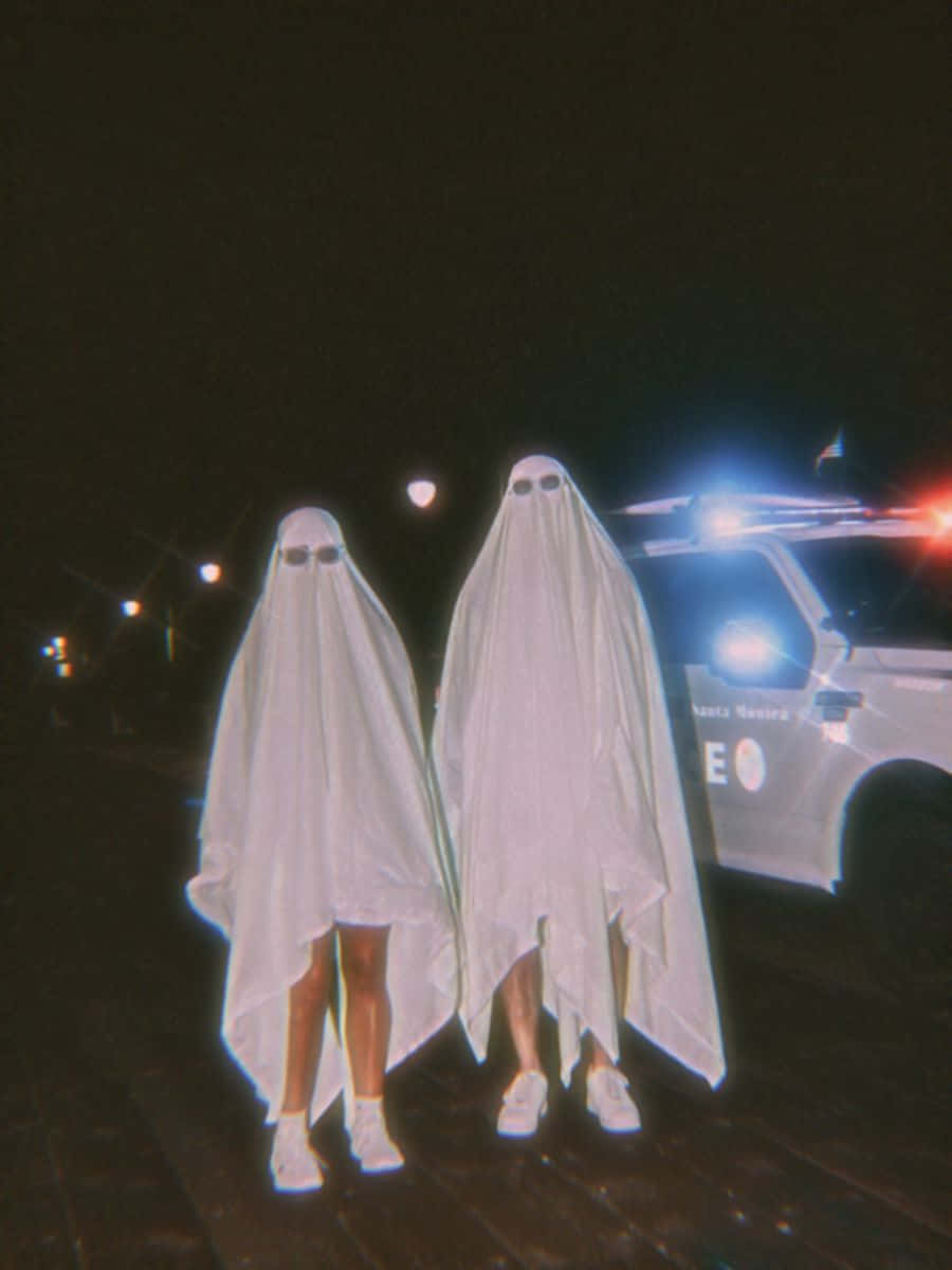 Baddie Vintage 90's Aesthetic 桌布 壁紙 — 兩個穿著白色鬼魂服裝的人站在一輛警車旁邊，夜間閃爍著燈光。
