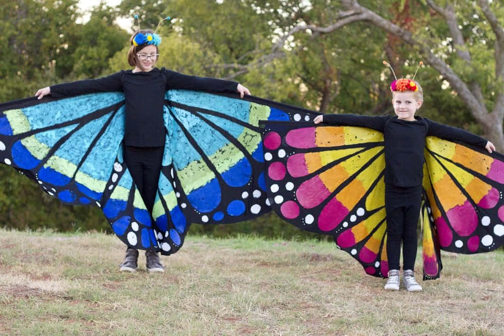 Butterfly Wing Dress 桌布 壁紙 — 兩個穿著色彩繽紛蝴蝶服裝的小孩在戶外樹下的草地上張開雙臂站立。