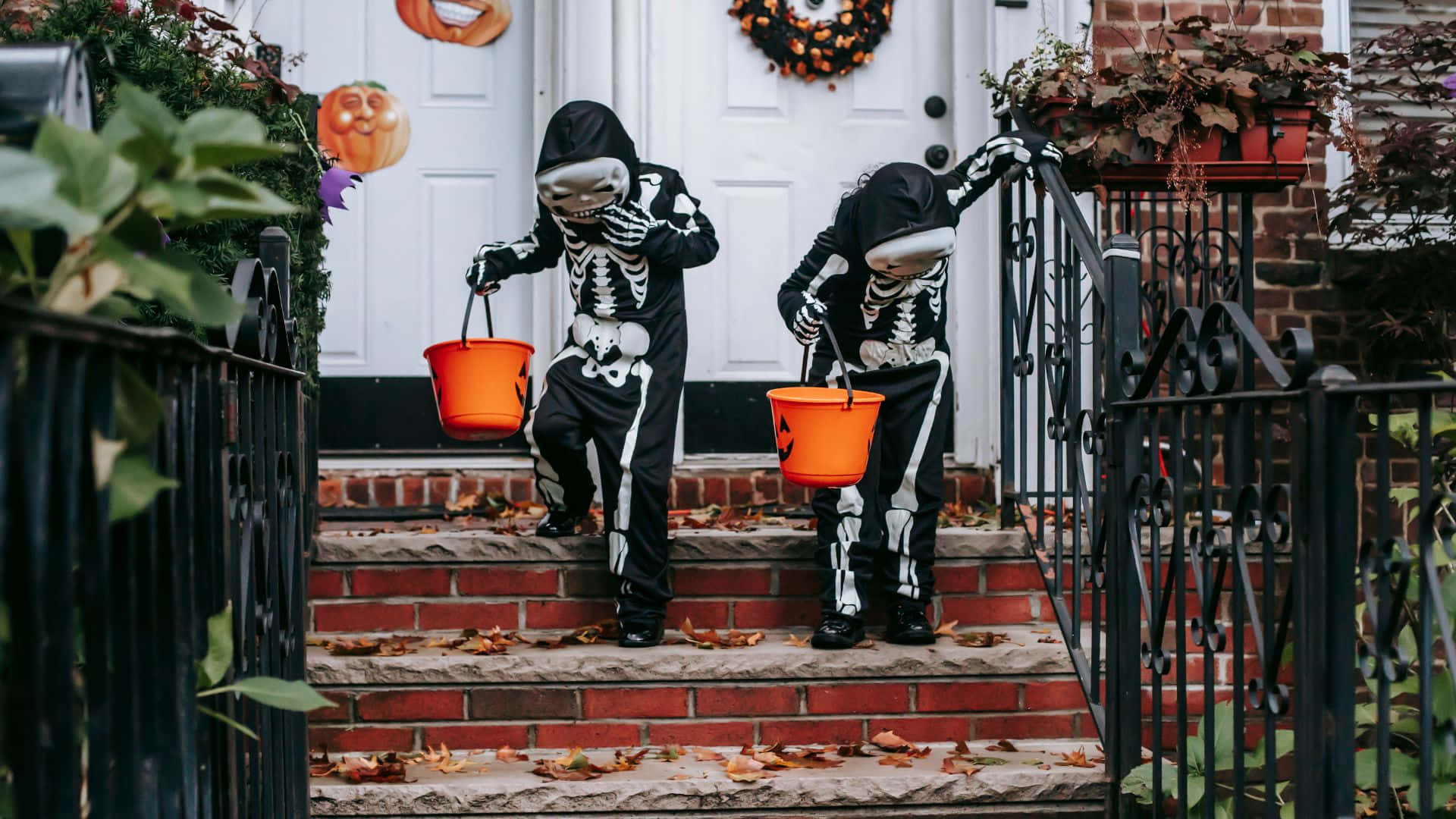 Halloween Costumes 桌布 壁紙 — 兩個穿著骷髏服裝、拿著橙色水桶的孩子爬上裝飾著秋葉和萬聖節裝飾的磚階。