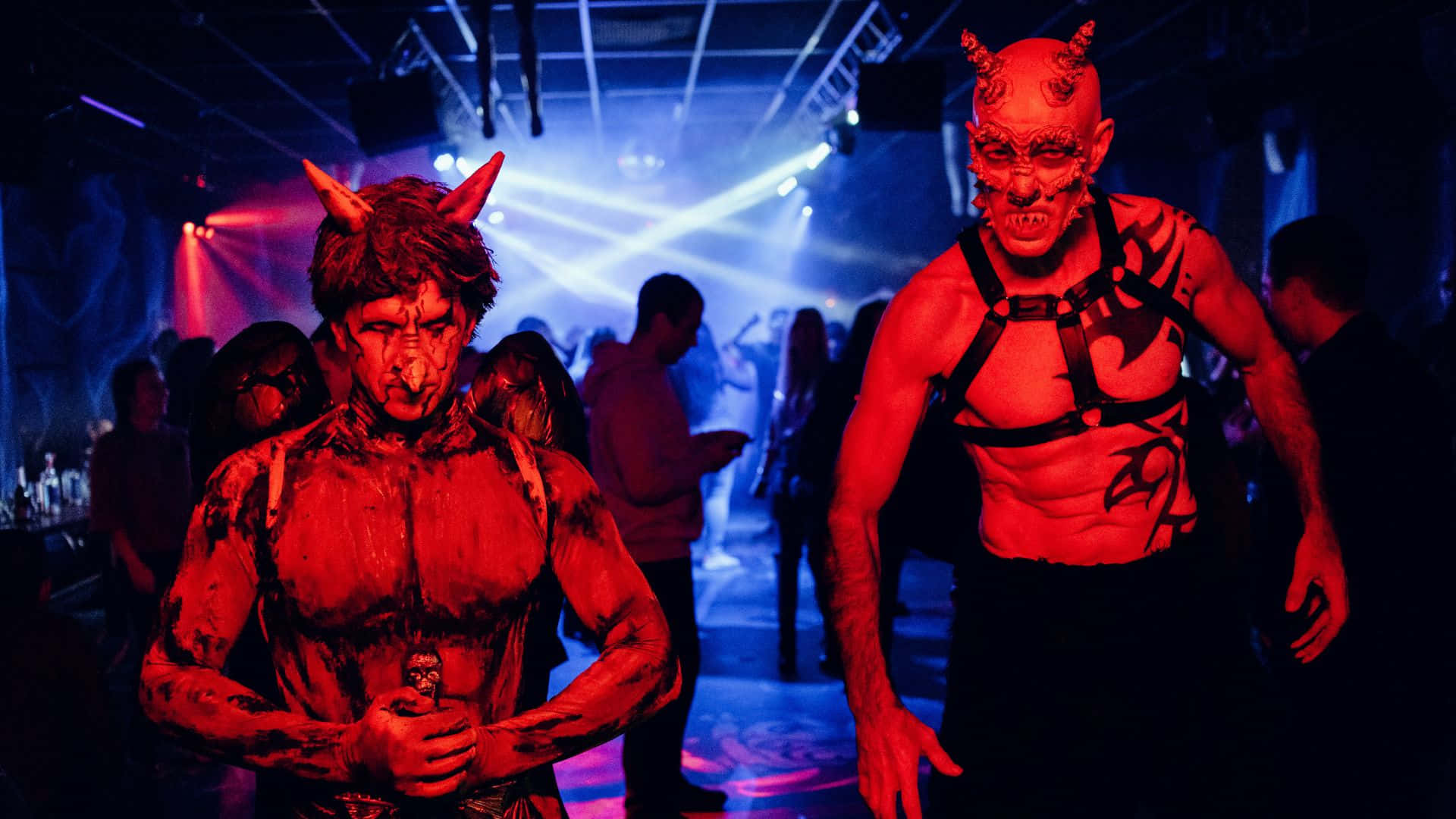 Horror Costumes 桌布 壁紙 — 兩個穿著魔鬼服裝、臉上塗著紅色顏料、頭上有角嘅男人企喺燈光昏暗嘅俱樂部入面，喺閃爍嘅藍色同紅色燈光下。