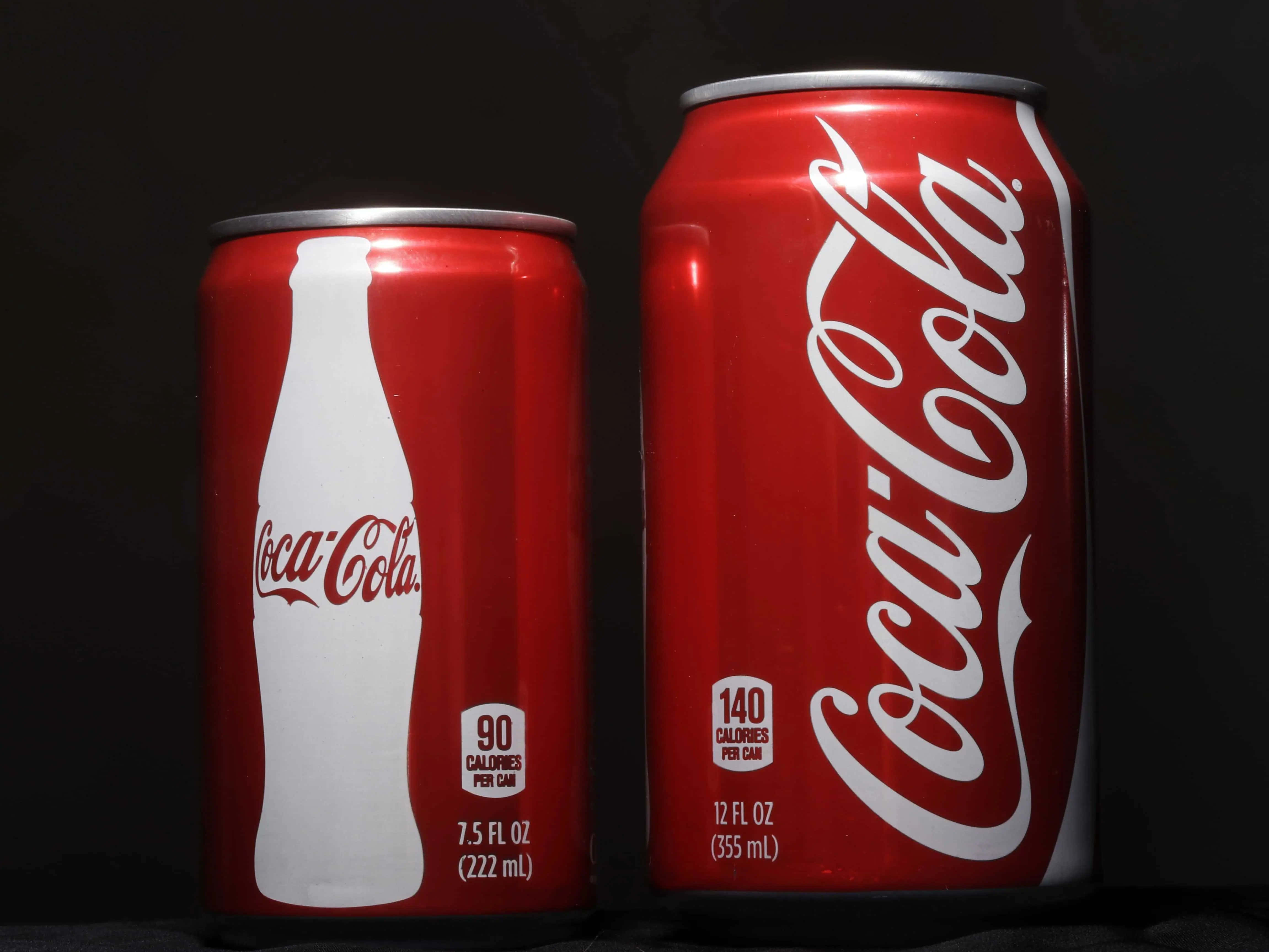 coca cola 桌布 壁紙 — 兩個紅色可口可樂罐，一個細罐有白色樽形標誌，另一個大罐有草書標誌，在深色背景前。