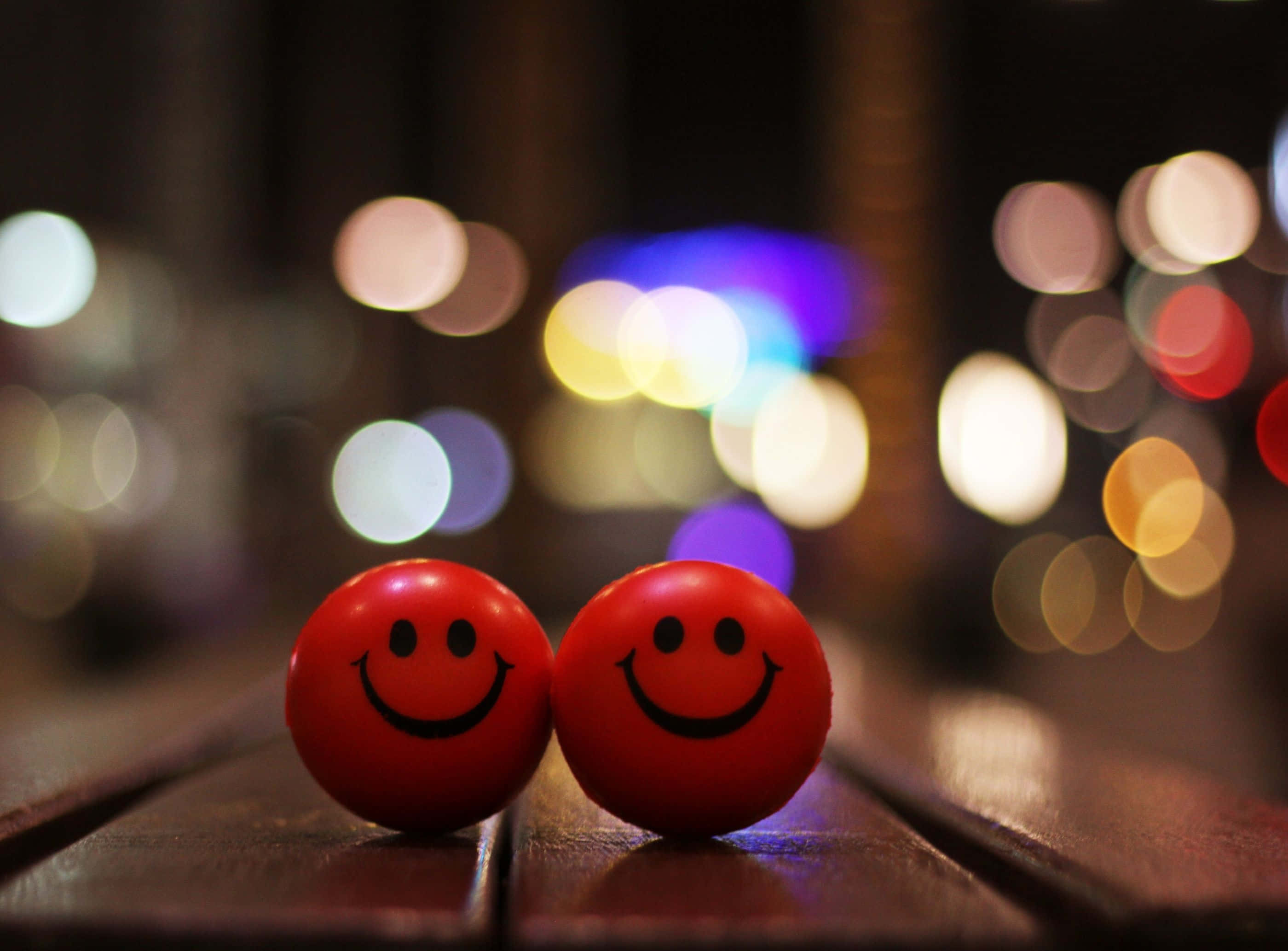 Cheerful 桌布 壁紙 — 兩個紅色笑臉波波並排放置在木質表面上，背景是充滿活力的散景城市夜景。