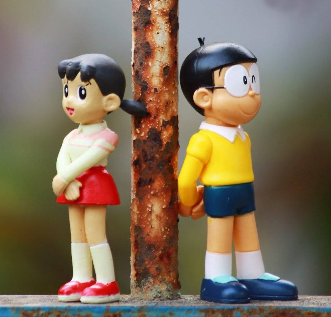 nobita shizuka love 桌布 壁紙 — 兩個經典動漫人偶背對背站在一根生鏽的柱子旁，背景是柔和模糊的戶外環境。