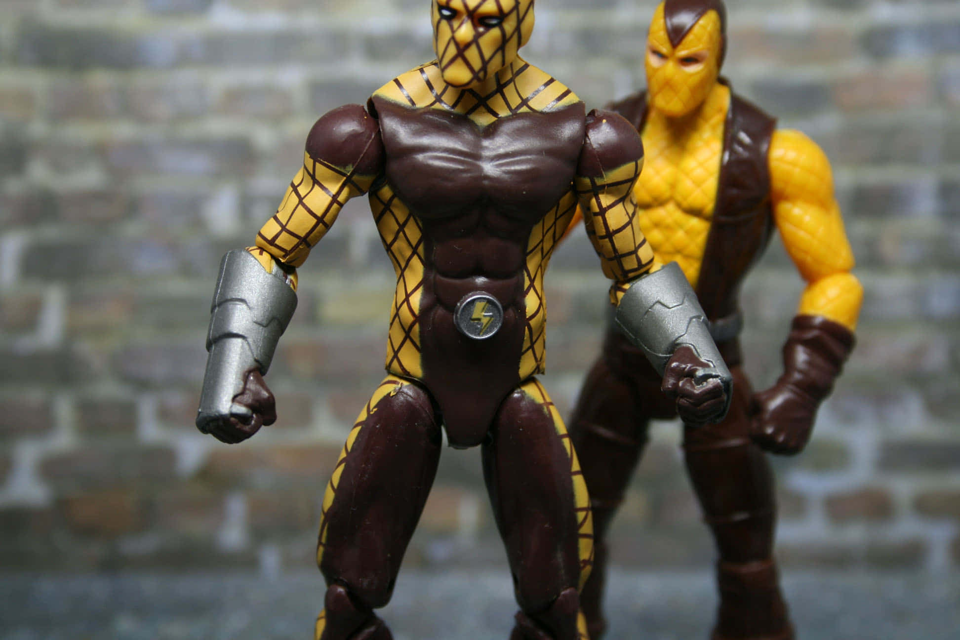 The Shocker 桌布 壁紙 — 兩個肌肉發達的動作人偶穿著黃色和棕色裝甲服，站在模糊的磚牆背景前。