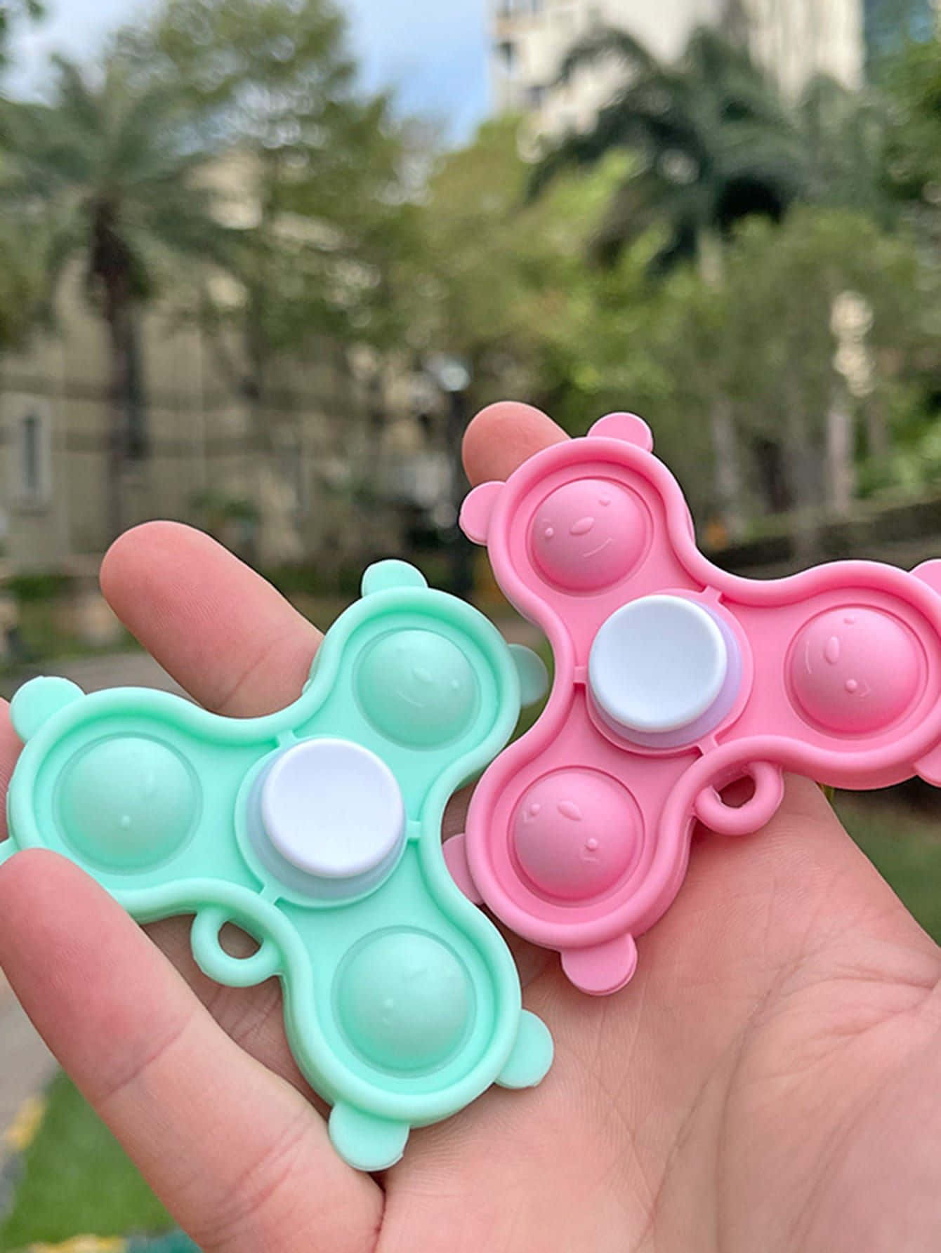 Popit 桌布 壁紙 — 兩個色彩繽紛的熊形按壓減壓玩具，握在手中，背景是模糊的戶外景色。