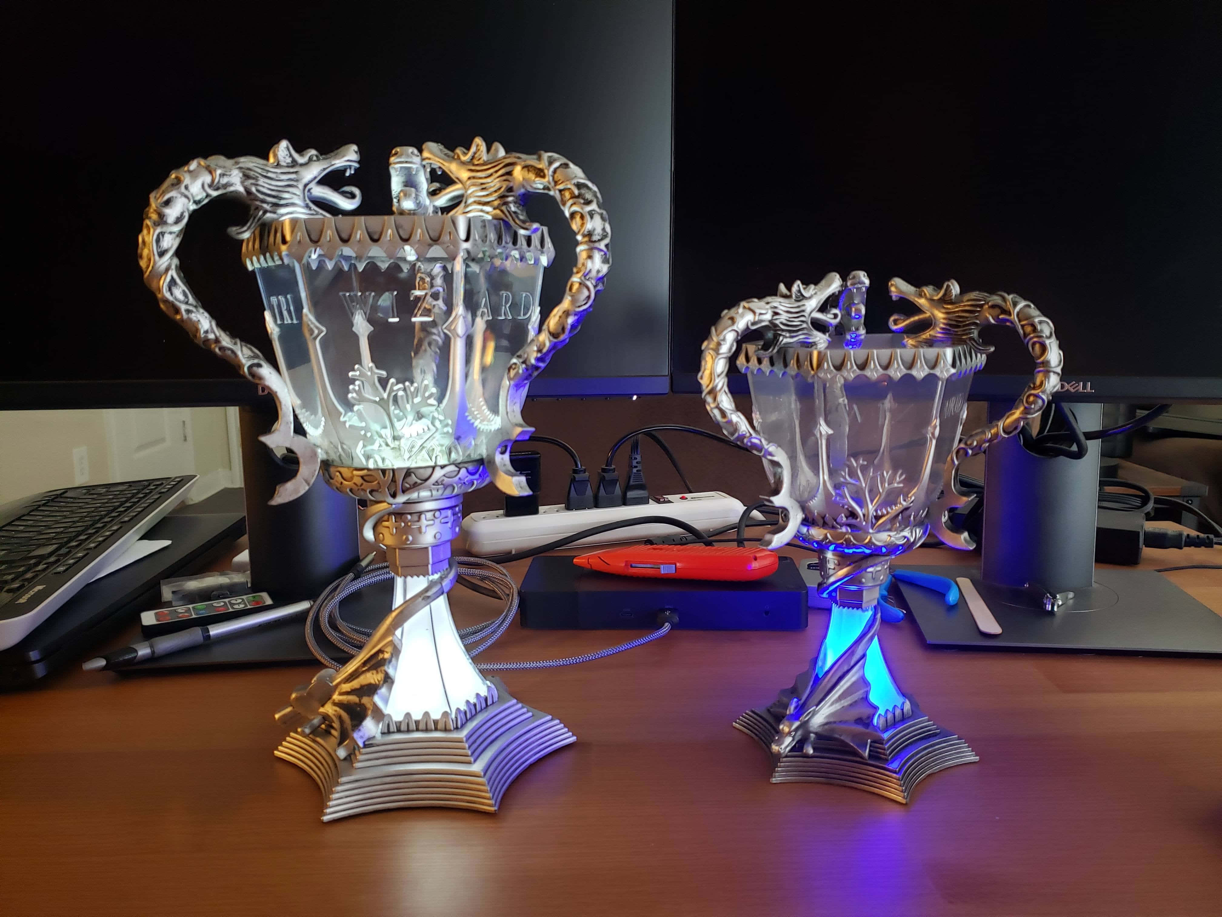 The Triwizard Cup 桌布 壁紙 — 兩個華麗的銀色獎盃，帶有龍形手柄，在雜亂的木桌上靠近顯示器發出藍光。