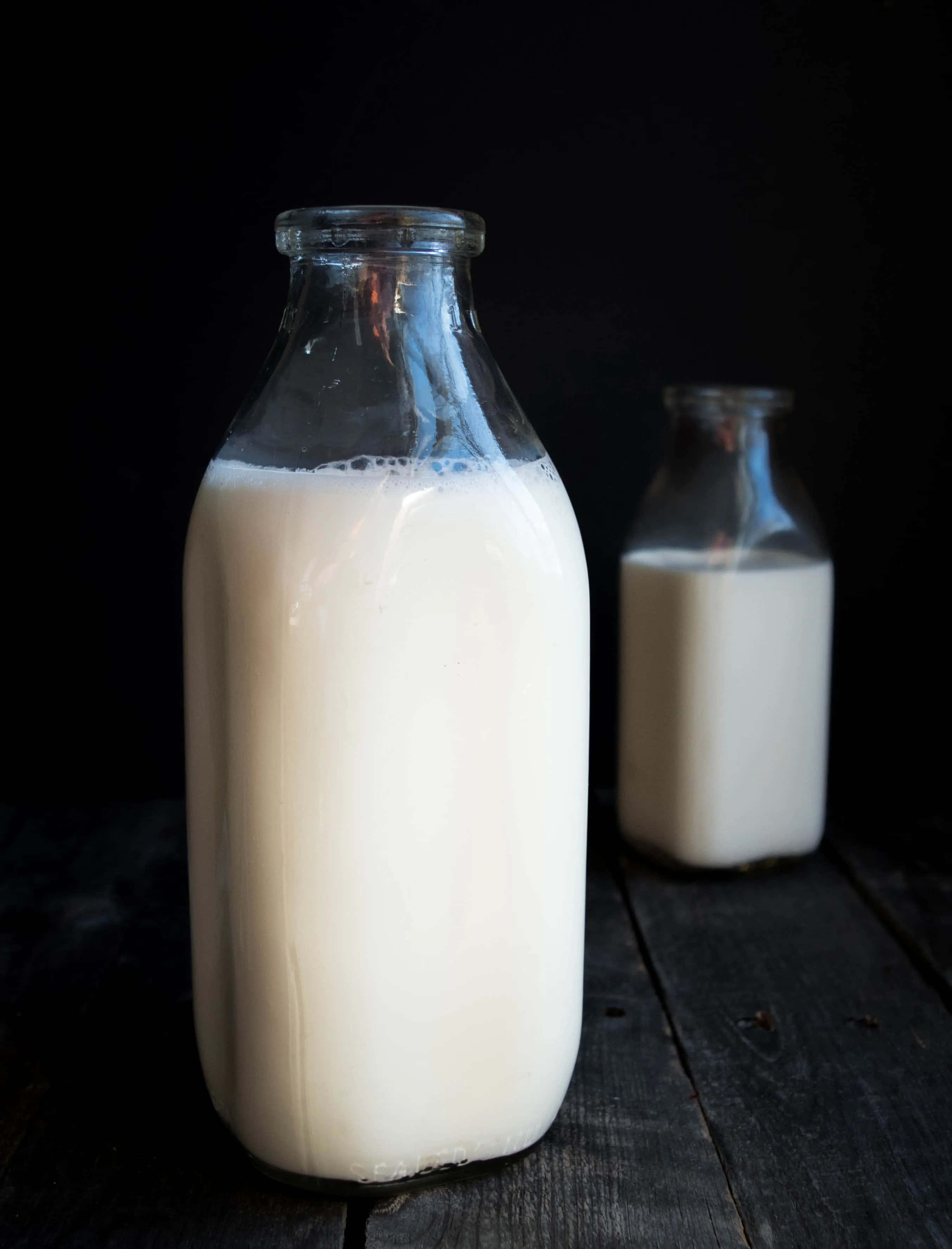 Milk 桌布 壁紙 — 兩個裝滿奶油白色牛奶的玻璃牛奶瓶放在黑色背景的深色木質表面上。