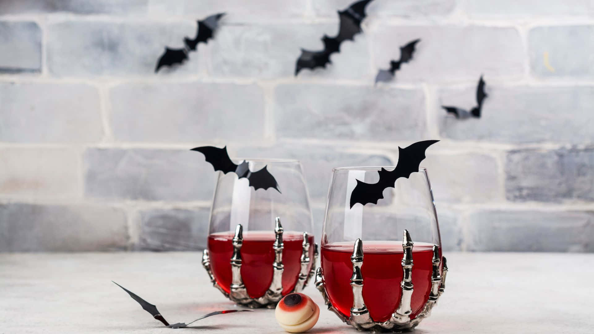 Spooky Cocktails 桌布 壁紙 — 兩個裝滿紅色液體的玻璃杯，裝飾著蝙蝠剪影和骷髏手，放在混凝土表面上，背景是灰色磚牆，上面有漂浮的蝙蝠裝飾。