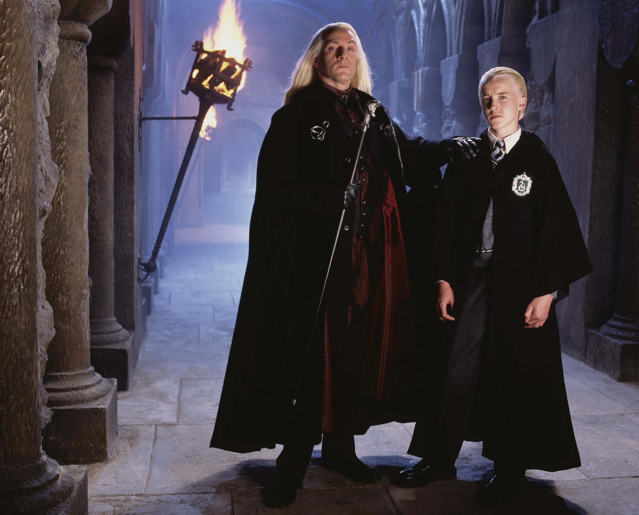 The Chamber Of Secrets 桌布 壁紙 — 兩個身穿黑色長袍的巫師站在一條由燃燒火炬照亮的石頭走廊中，投射出戲劇性的陰影。