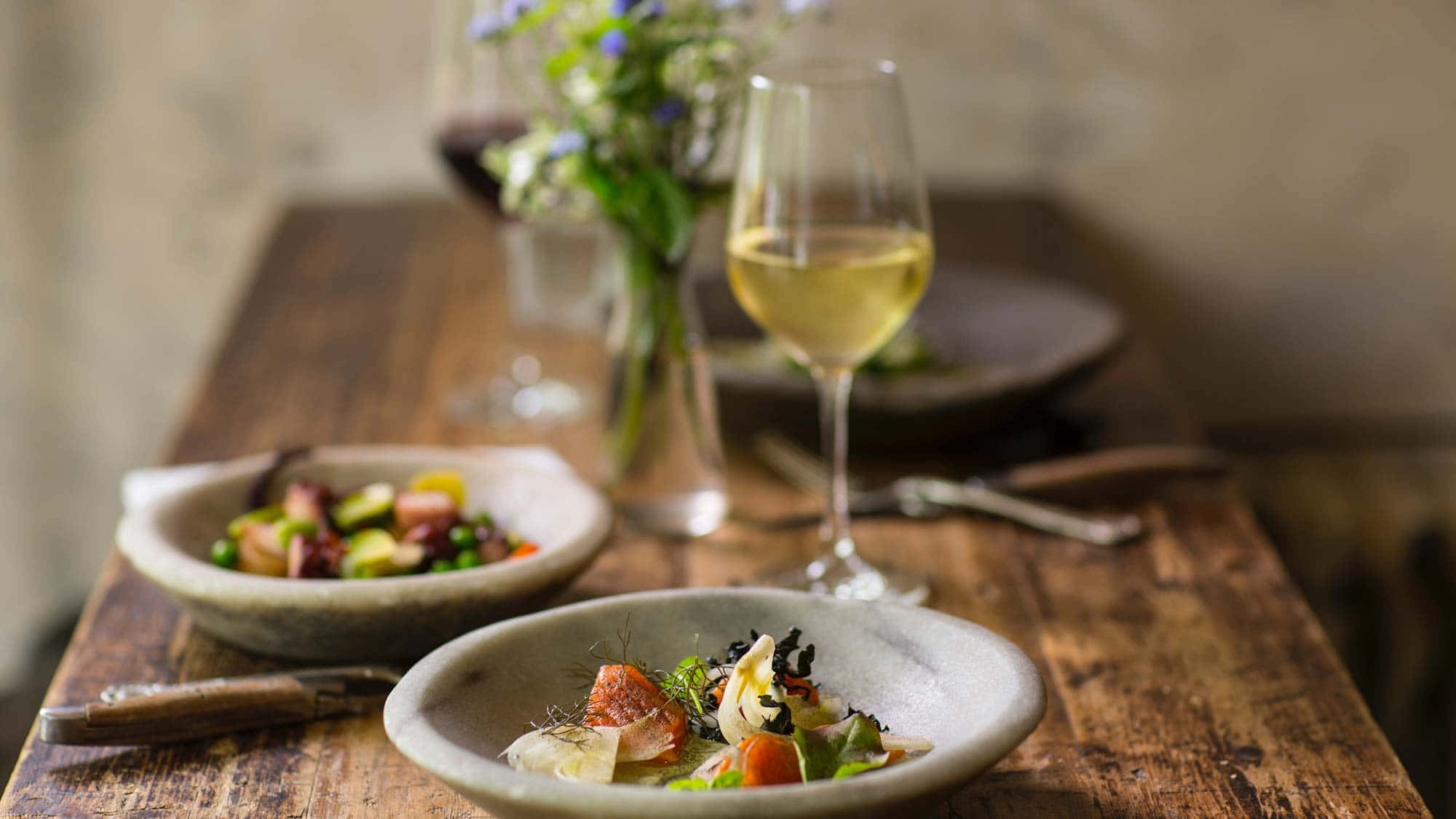 Food And Beverage 桌布 壁紙 — 兩個鄉村風格的碗盛載色彩繽紛的美食沙律，放在一張經風化的木桌上，旁邊有白酒和紅酒杯，以及一束藍色花朵的花瓶，背景柔和。