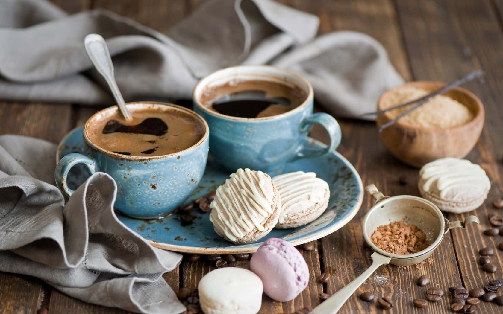 Winter Desserts 桌布 壁紙 — 兩個鄉村風格的藍色咖啡杯，搭配馬卡龍和散落的咖啡豆，放在鋪有灰色布的木桌上。
