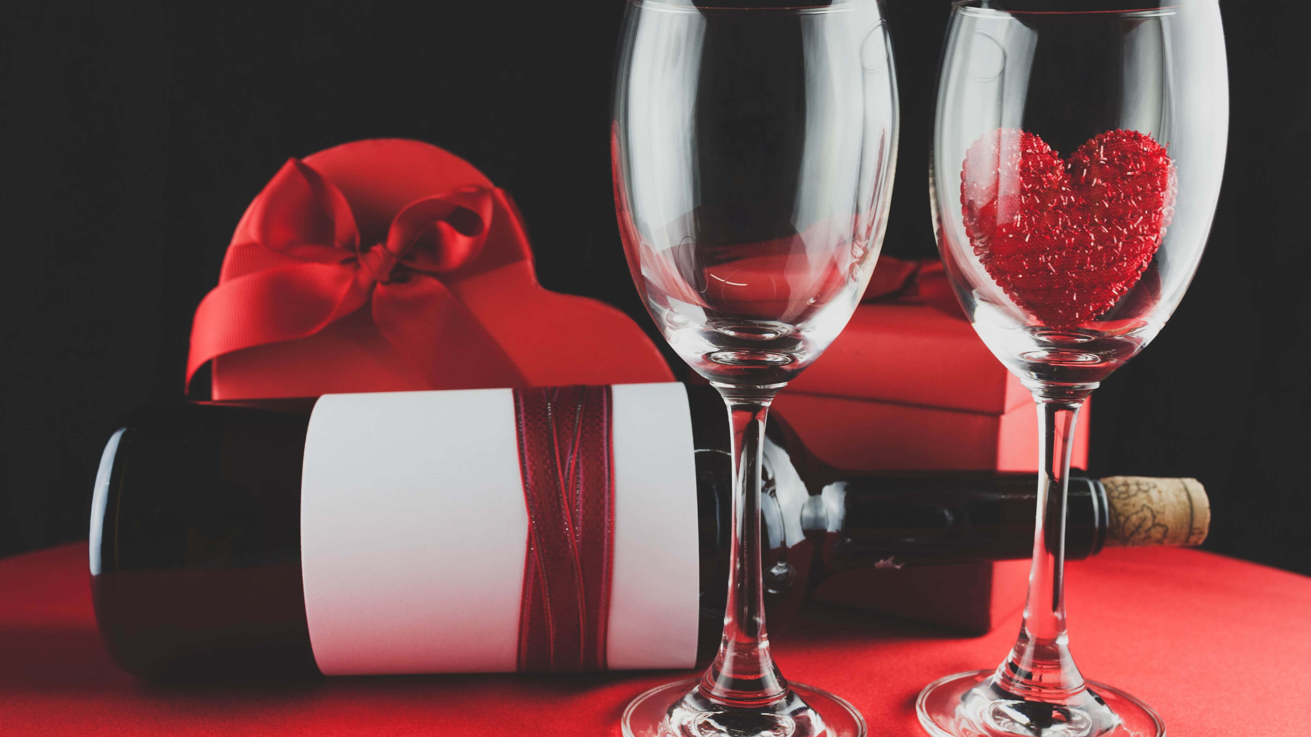 Red Wine Glass 桌布 壁紙 — 兩個酒杯，其中一個裝著閃閃發光的紅色心形，旁邊放著一個酒瓶和一個紅色禮物盒，放在紅色表面上。