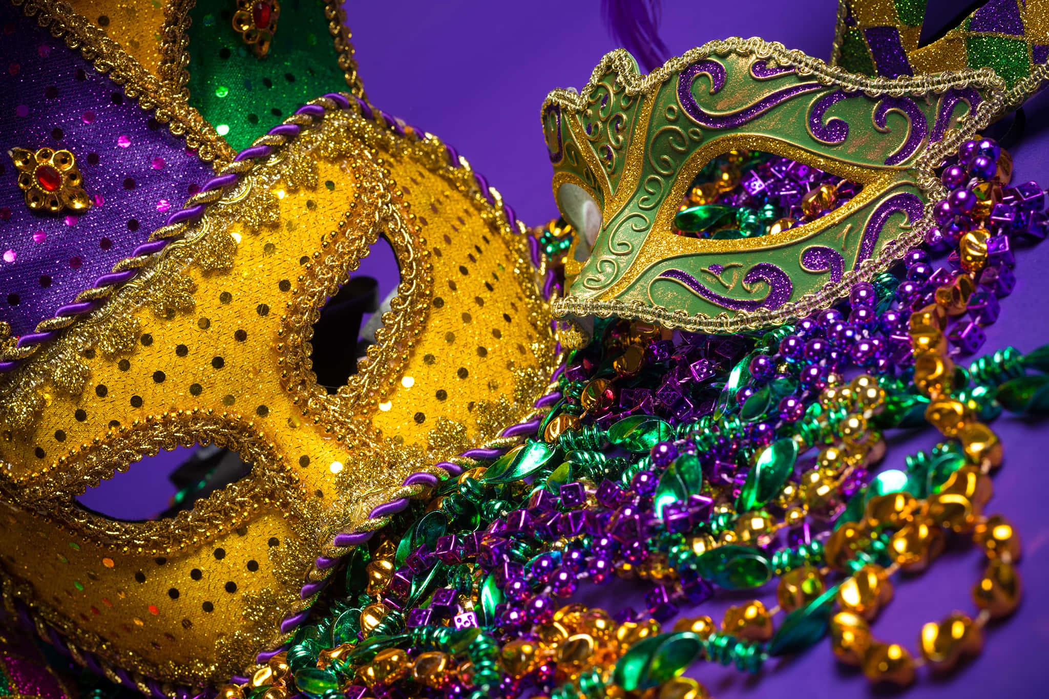 Mardi Gras 桌布 壁紙 — 兩個金色、紫色和綠色閃粉裝飾的狂歡節面具放在一堆相配的珠子上，背景是深紫色。