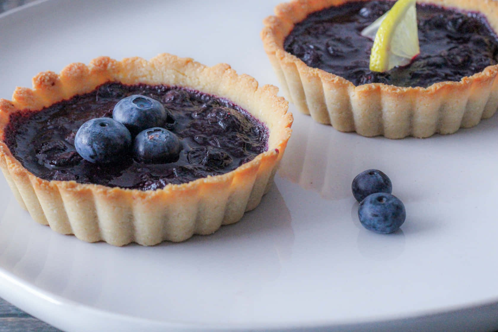 Blueberries Tart 桌布 壁紙 — 兩個金色藍莓撻，帶有光澤的餡料，上面放上新鮮漿果和檸檬角，放在白色盤子上。