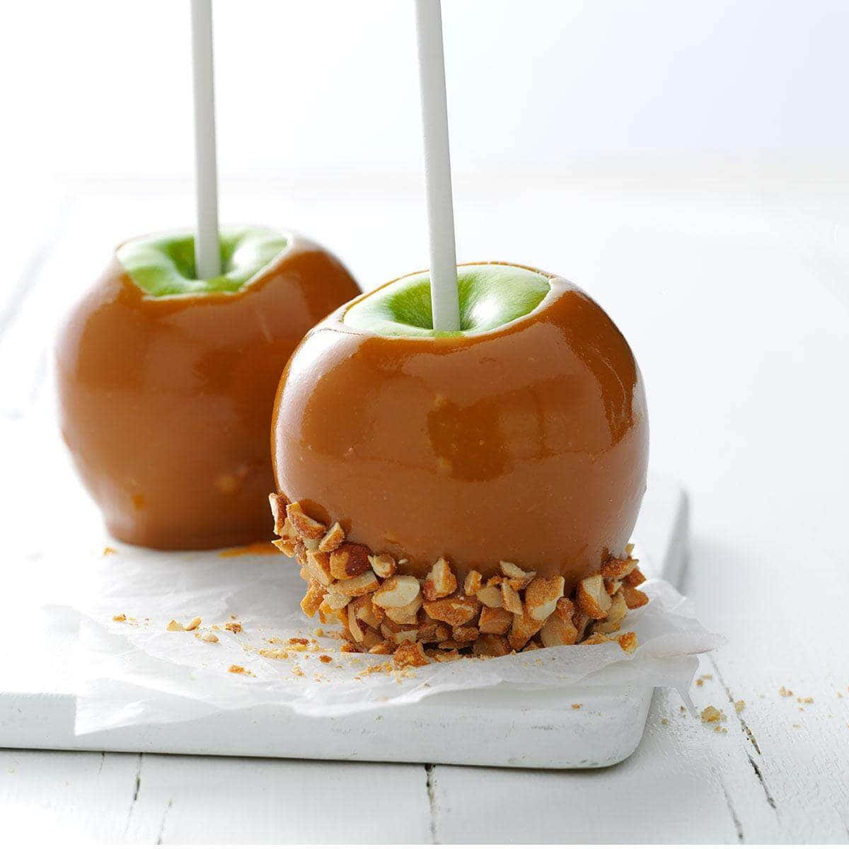 Caramel Apples 桌布 壁紙 — 兩個閃亮的焦糖蘋果，上面有切碎的堅果，放在白色木板上，背景明亮。