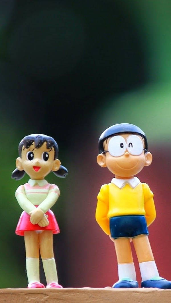 Doraemon 桌布 壁紙 — 兩個開心的動漫公仔多啦A夢和靜香並排站在木質表面上，背景是模糊的綠色和紅色。