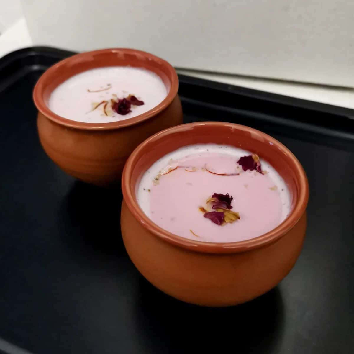 Pink Tea 桌布 壁紙 — 兩個陶土花盆裝滿粉紅色和白色的牛奶飲料，上面撒有玫瑰花瓣和藏紅花絲，放在黑色托盤上。