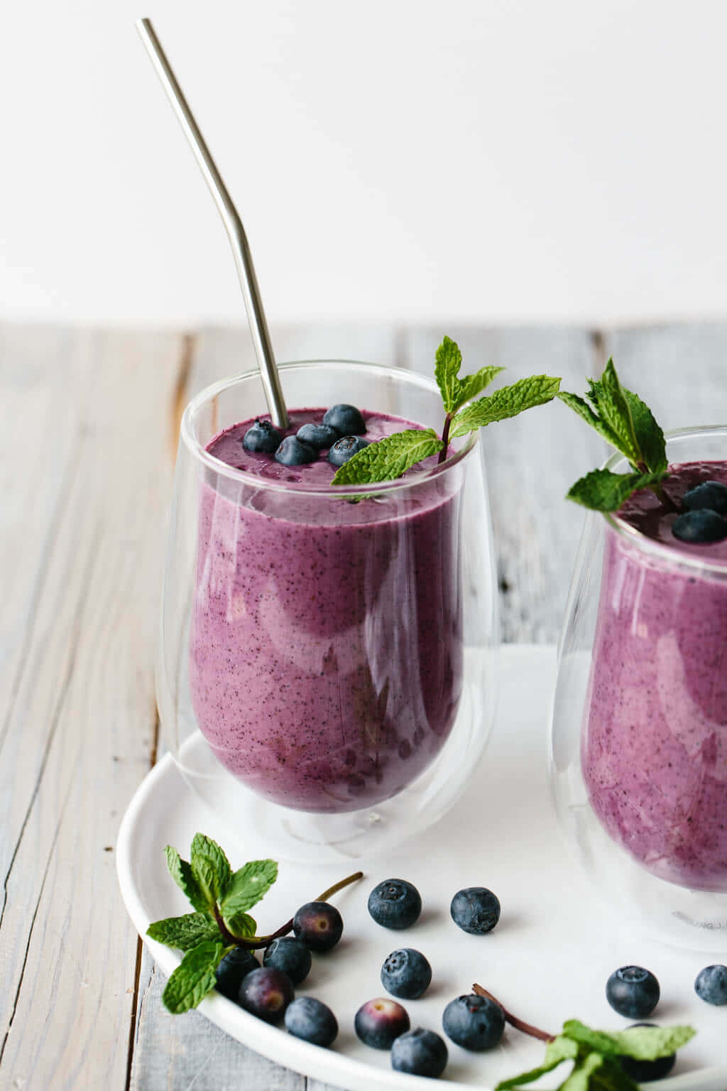 Blueberry Smoothie 桌布 壁紙 — 兩個雙層玻璃杯中的鮮豔紫色藍莓冰沙，飾以新鮮薄荷和整顆藍莓，放在白色碟子上。