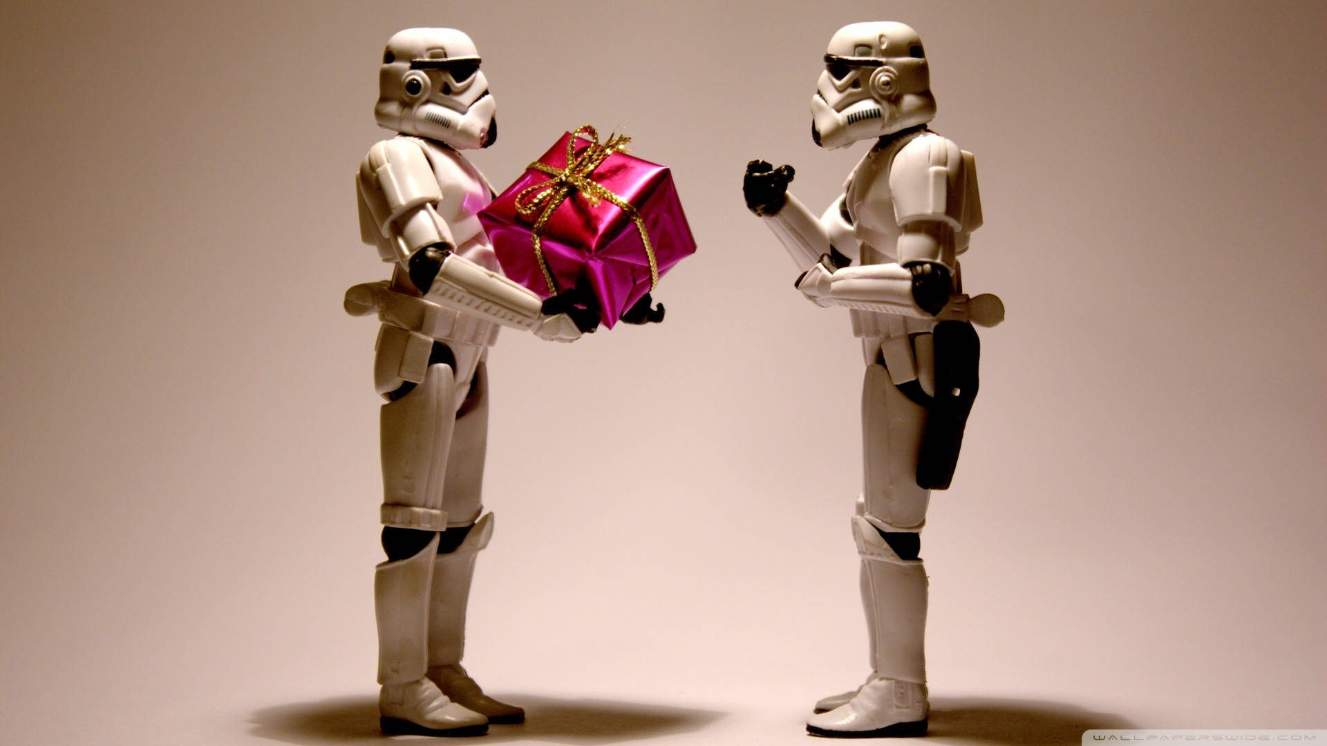 Stormtrooper 桌布 壁紙 — 兩個風暴兵人偶在純米色背景前交換一個閃亮的粉紅色禮物，禮物用金色絲帶包裝。