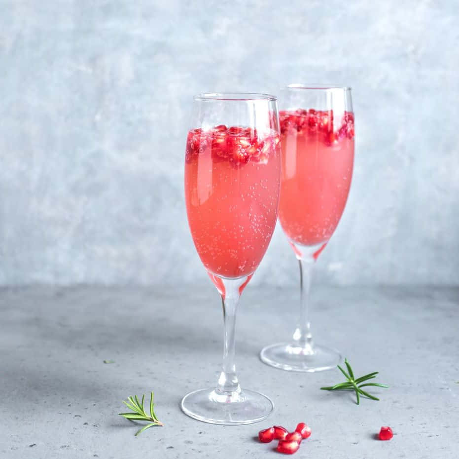 Pink Champagne 桌布 壁紙 — 兩個香檳杯盛滿閃爍粉紅石榴飲品，飾以新鮮迷迭香和散落的石榴籽，置於灰色表面上。