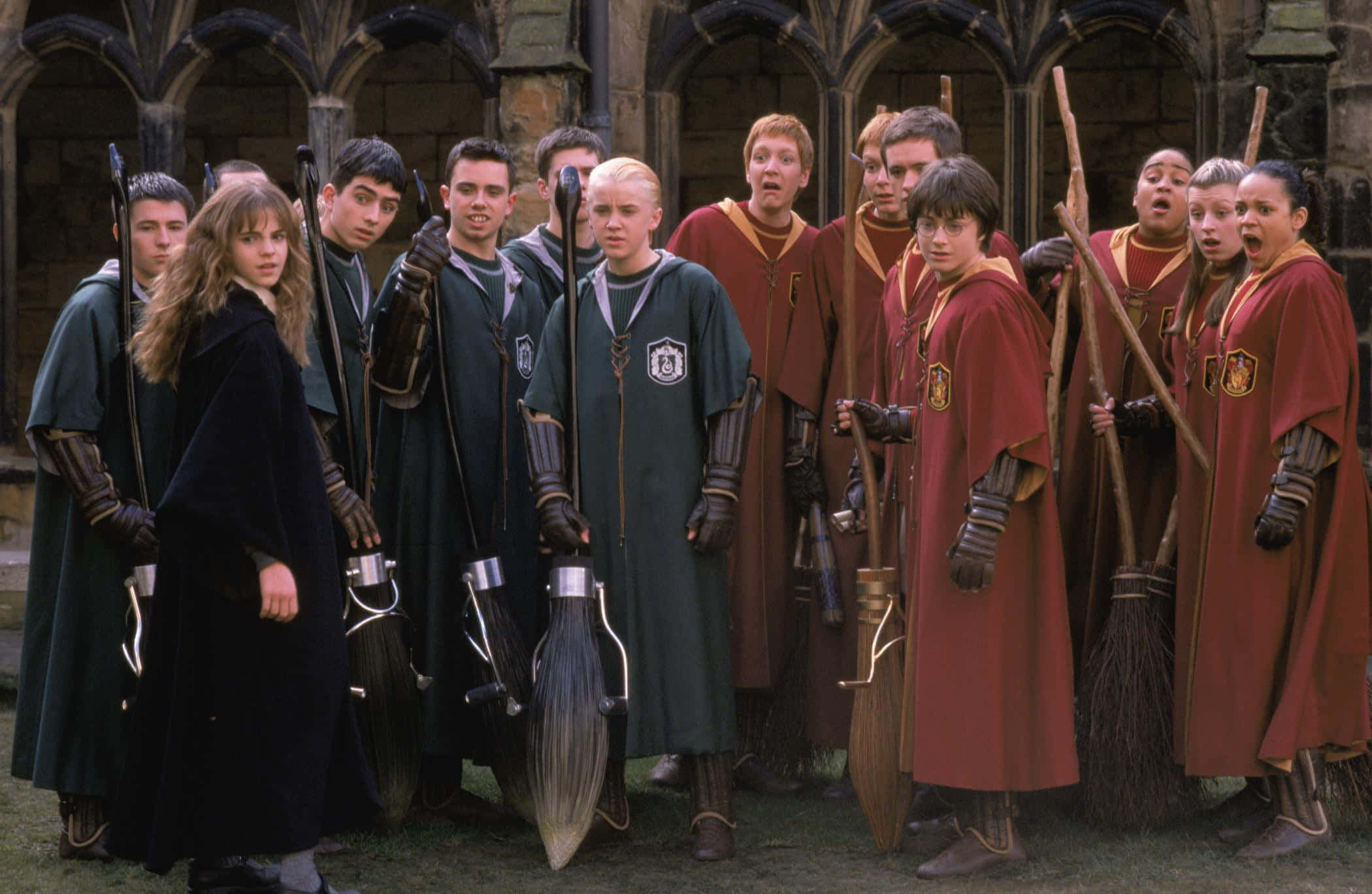 The Chamber Of Secrets 桌布 壁紙 — 兩個魁地奇隊伍穿著綠色和紅色長袍，在石頭庭院中手持掃帚，表情緊張而專注。
