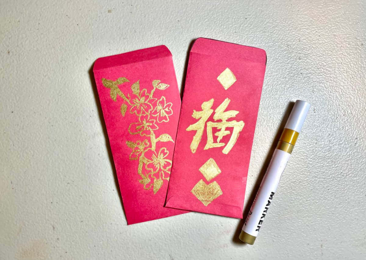 Red Envelope 桌布 壁紙 — 兩個鮮紅色嘅利是封，上面有金色花卉同福字，旁邊放咗一支白色記號筆，喺有紋理嘅米白色表面上。