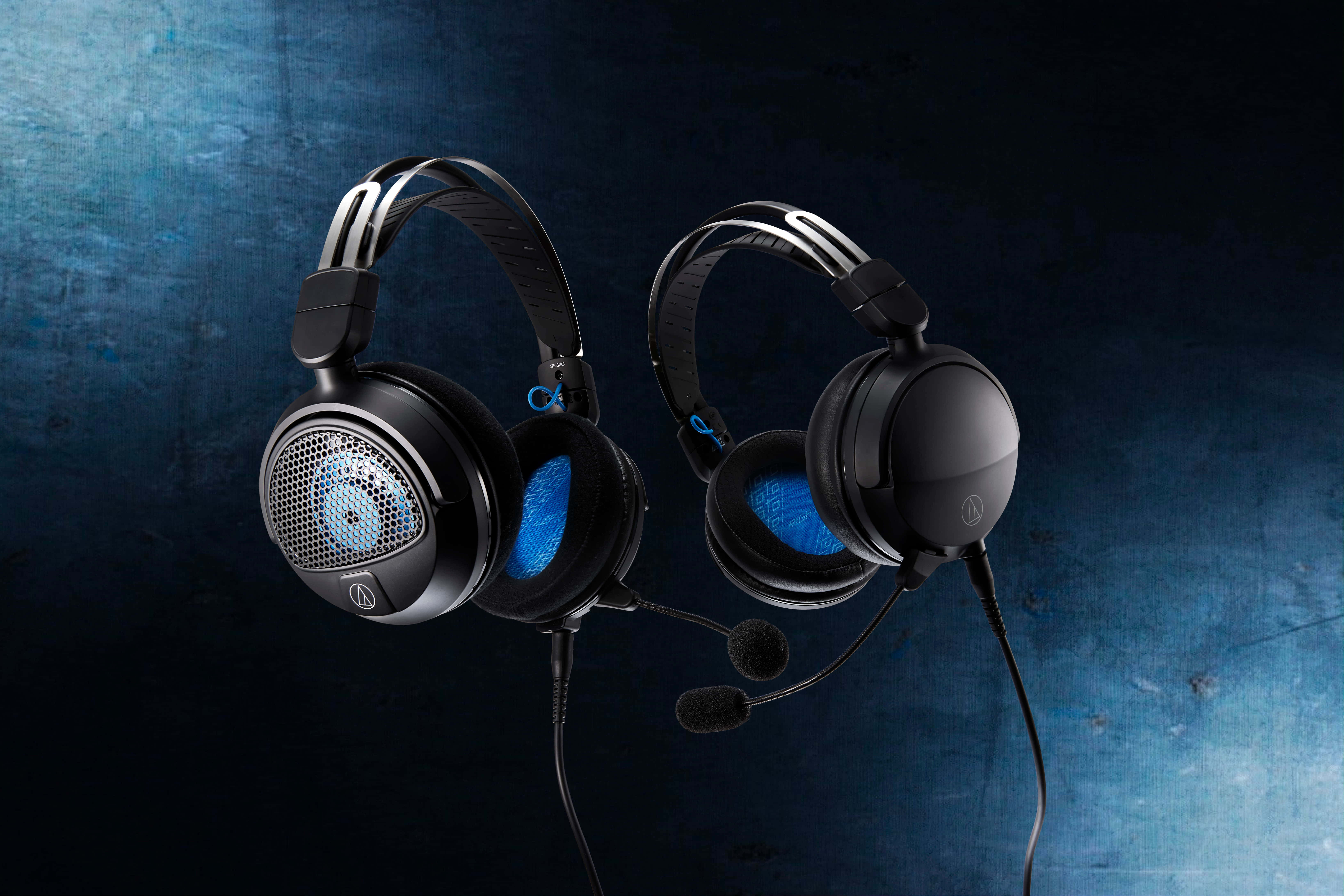 Gaming Headsets 桌布 壁紙 — 兩個黑色遊戲耳機，帶有藍色發光耳罩和麥克風，懸掛在帶有紋理的深藍色背景上。