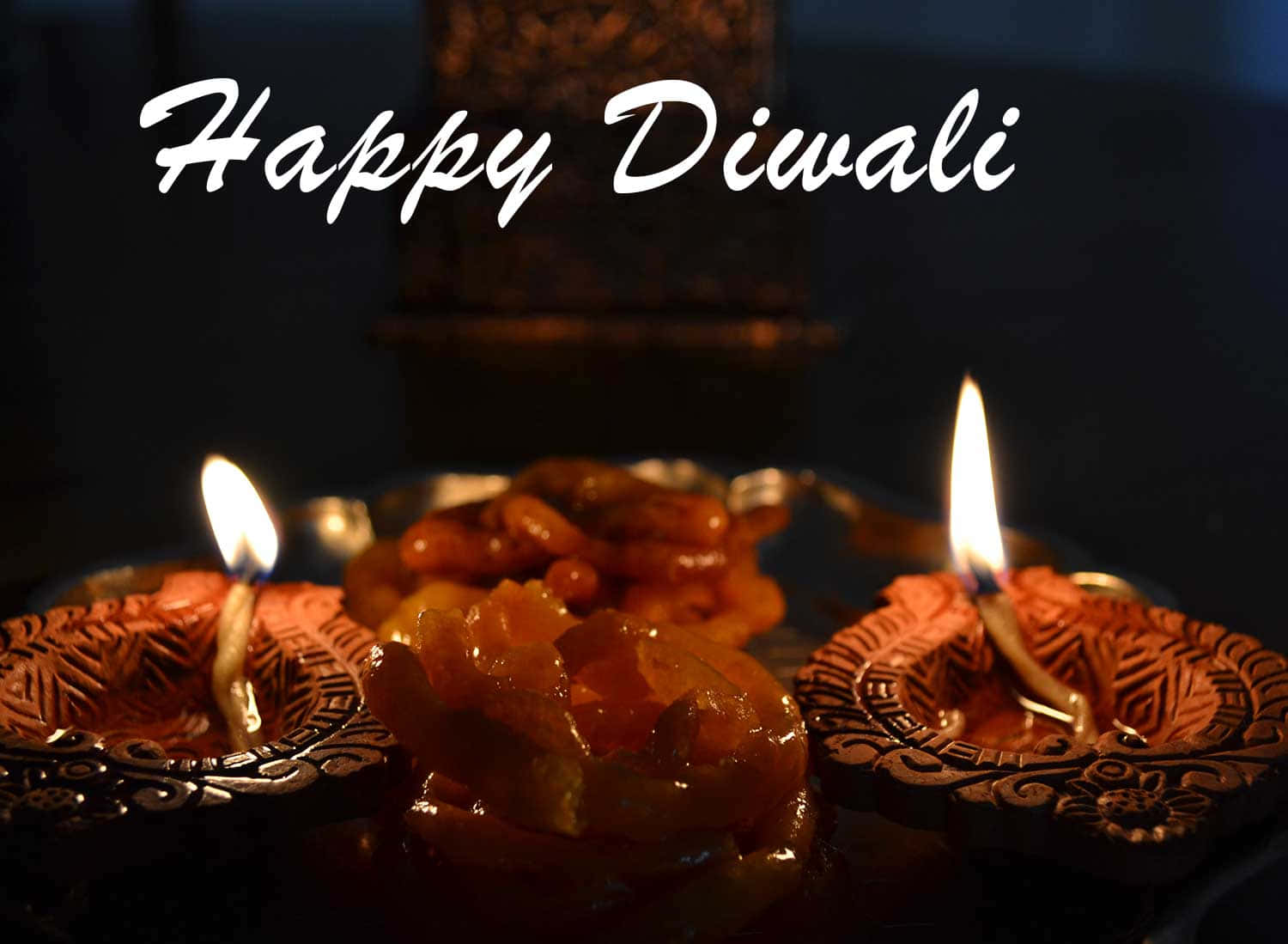 Happy Diwali 桌布 壁紙 — 兩個點燃的黏土迪亞燈在黑暗背景中，旁邊有一盤甜點，並帶有「Happy Diwali」文字，溫暖地發光。