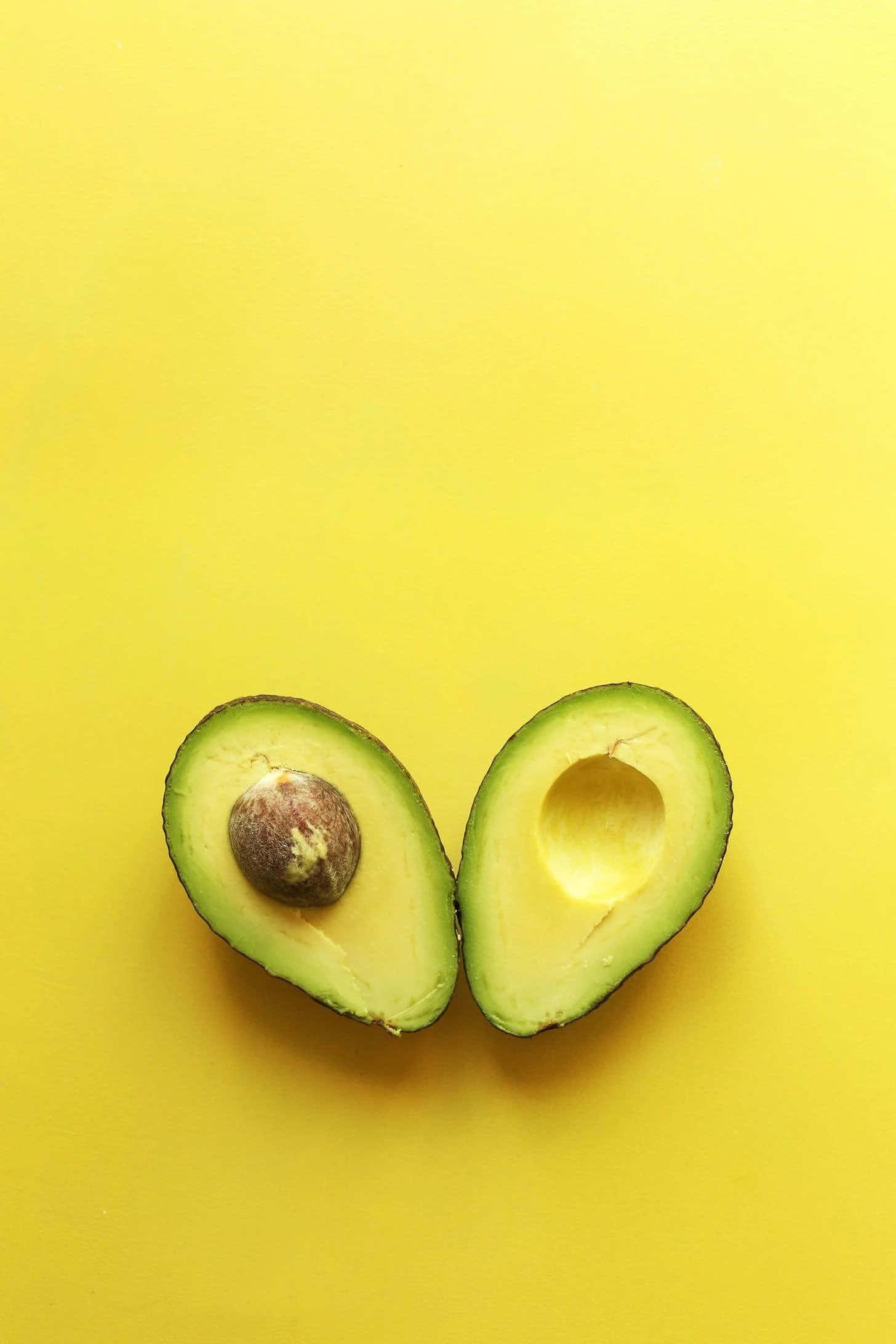 Avocado 桌布 壁紙 — 兩半成熟的牛油果連核放在鮮黃色背景上。