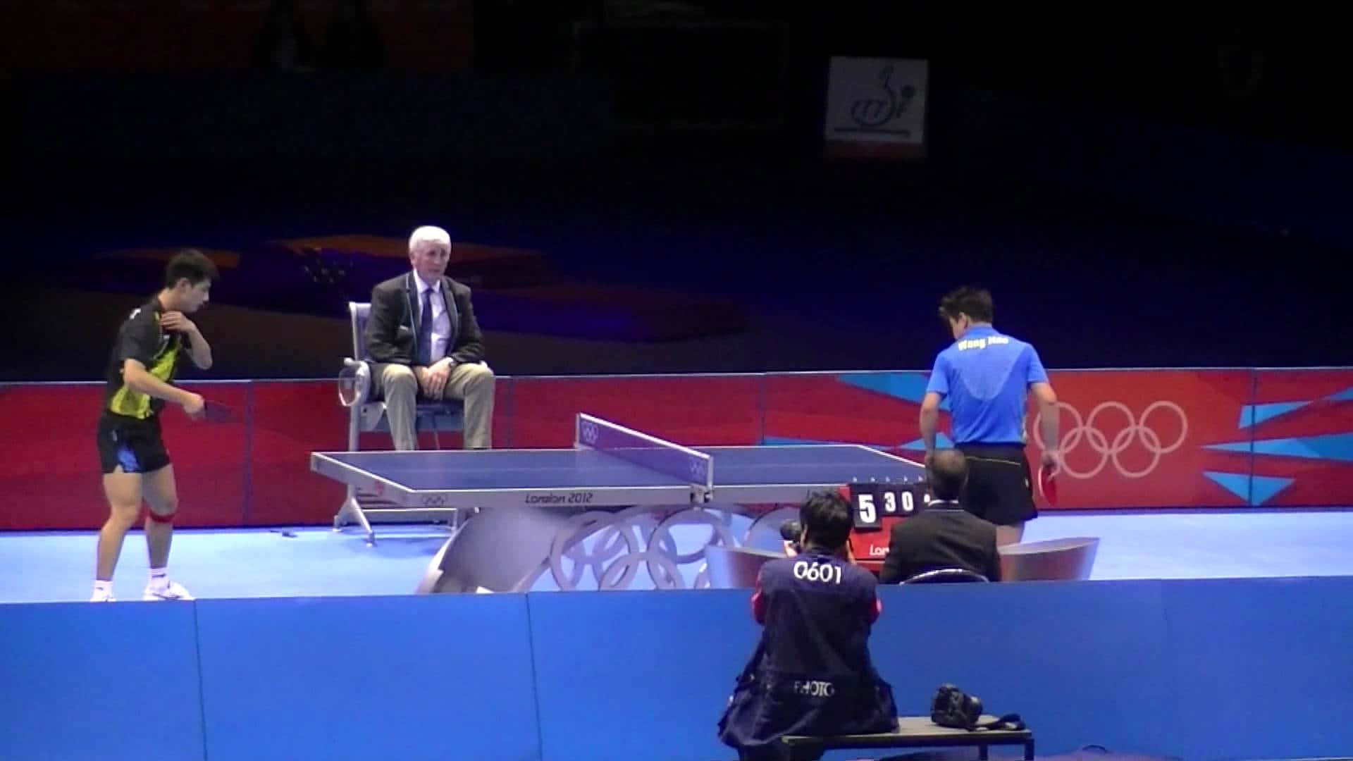Table Tennis 桌布 壁紙 — 兩名乒乓球運動員在明亮的場館燈光下比賽，一名裁判坐在網邊，奧運品牌標誌清晰可見。