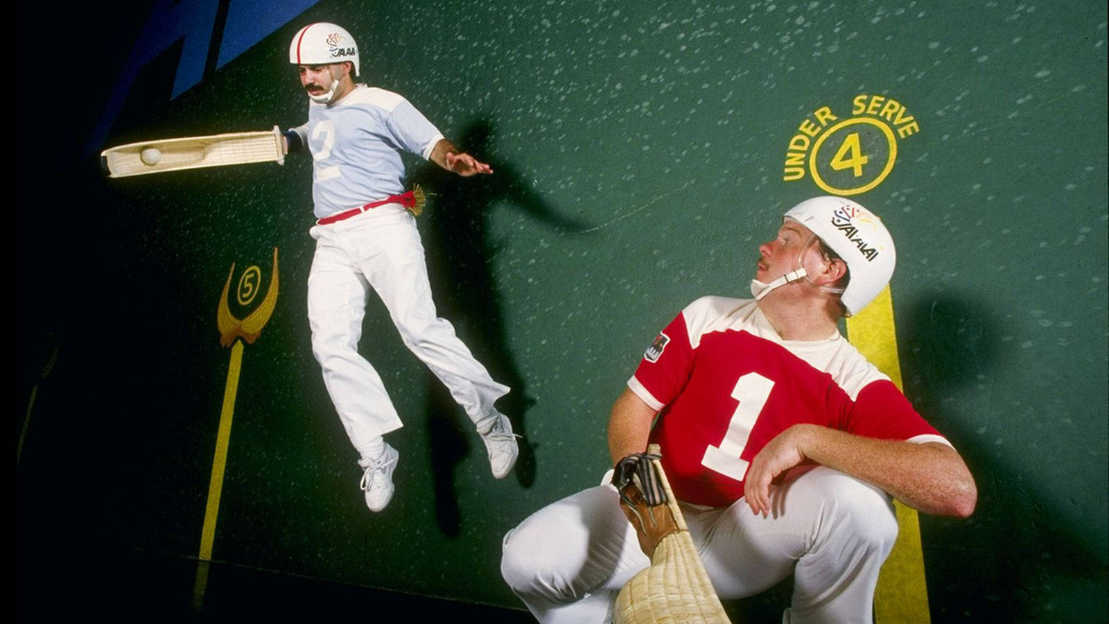 Jai Alai Blues 壁紙 — 兩名回力球運動員正在比賽，一名在空中手持cesta，另一名靠著帶有黃色標記的綠色牆壁坐著。