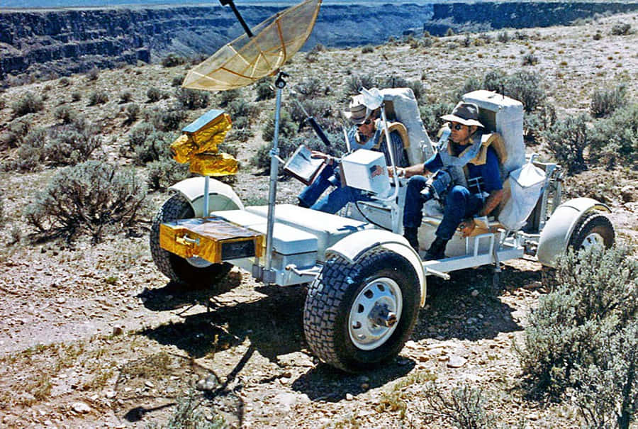 Lunar Rover 桌布 壁紙 — 兩名太空人乘坐月球車原型，穿過陽光明媚的沙漠峽谷，地形多岩石，灌木稀疏。
