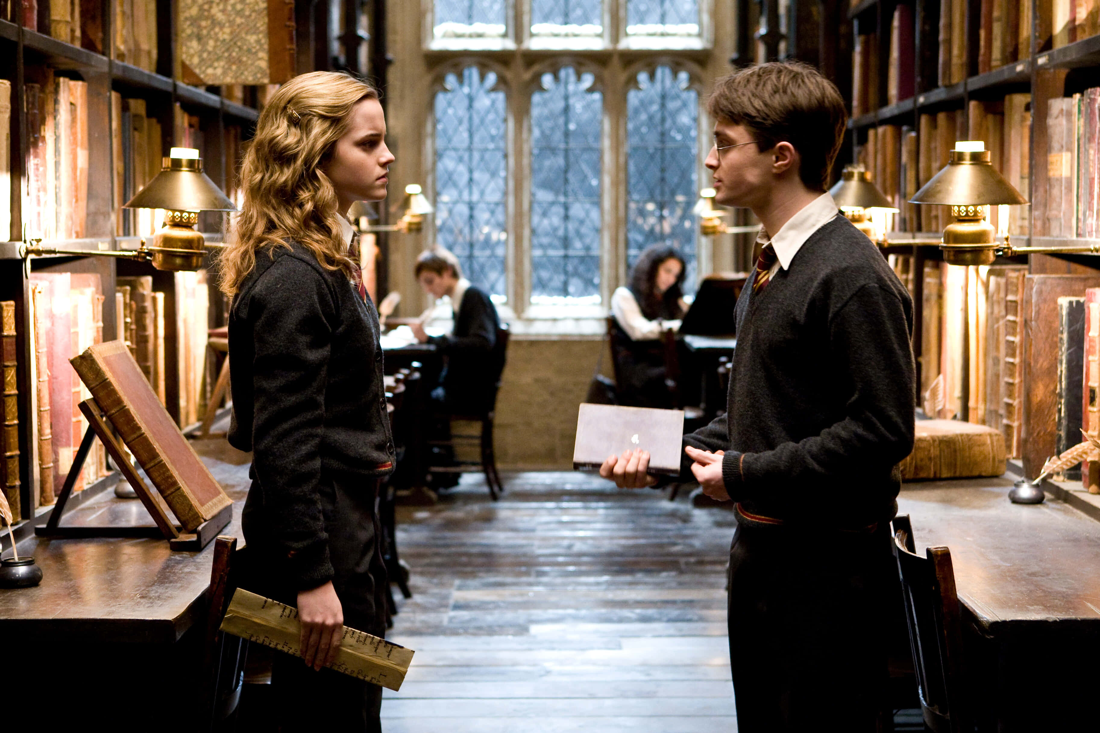 The Hogwarts Library 桌布 壁紙 — 兩名學生在一個宏偉、燭光映照的圖書館中對峙，館內佈滿高聳的書架和彩繪玻璃窗。